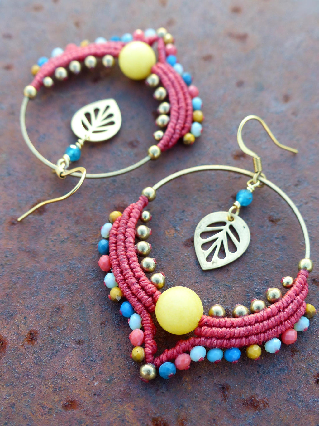 Peut inclure: Une paire de boucles d'oreilles créoles avec du macramé rouge et or, des perles jaunes et des perles bleues et blanches. Les boucles d'oreilles ont un charme en forme de feuille d'or suspendu.