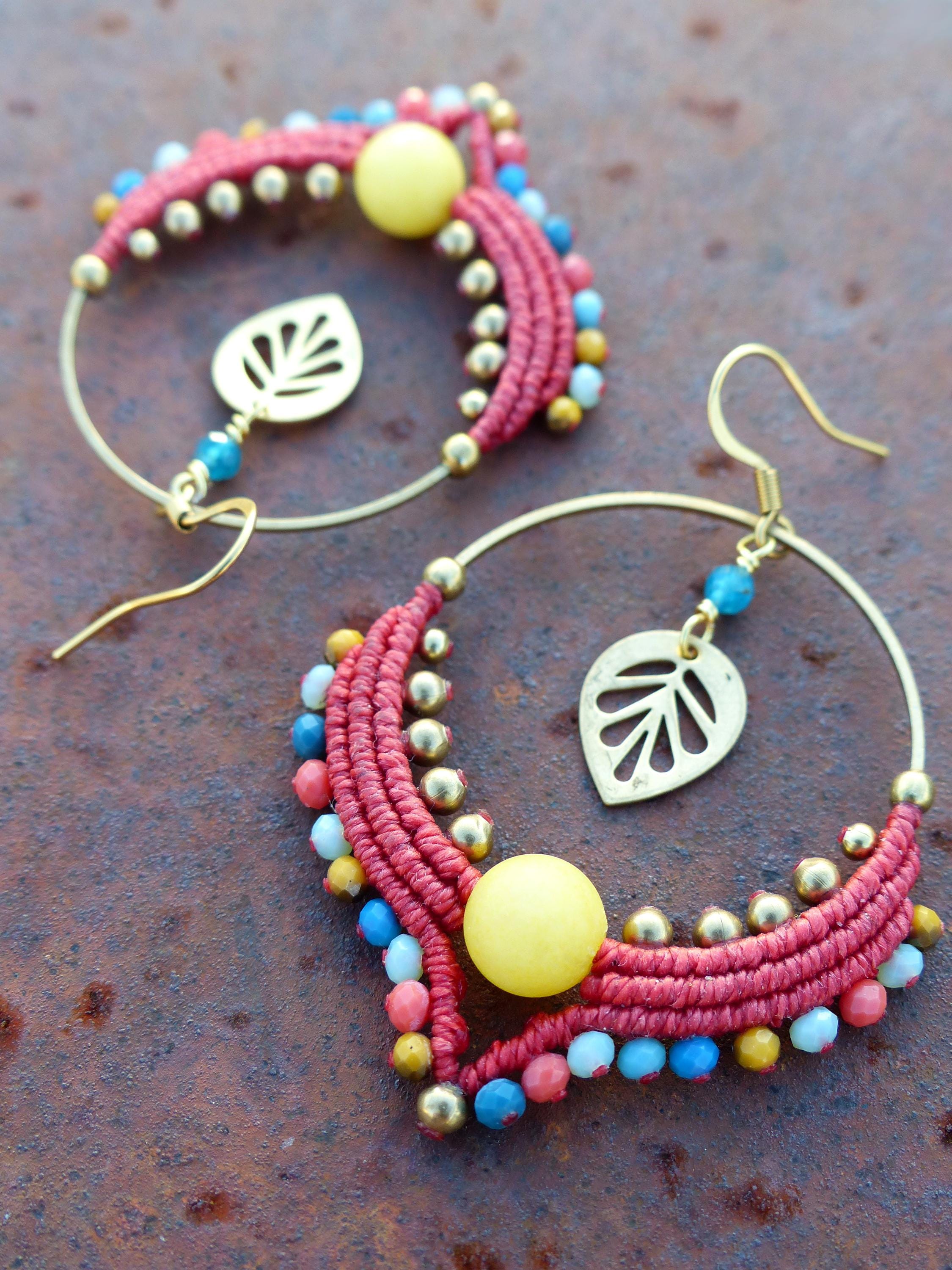 Peut inclure: Une paire de boucles d'oreilles créoles avec du macramé rouge et or, des perles jaunes et des perles bleues et blanches. Les boucles d'oreilles ont un charme en forme de feuille d'or suspendu.