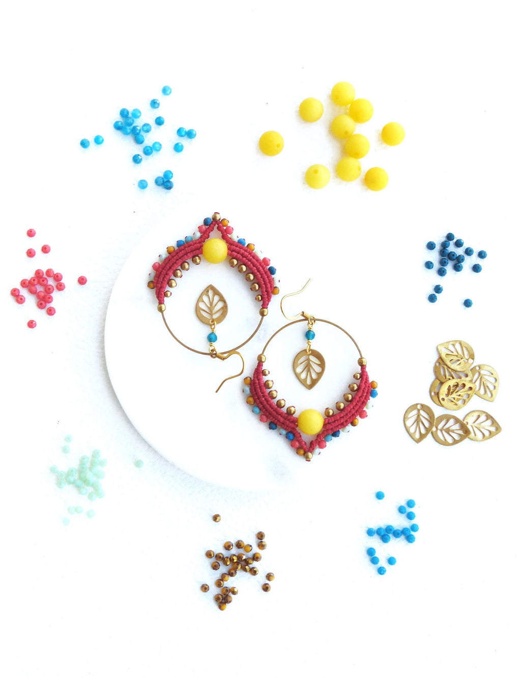 Peut inclure: Une paire de boucles d'oreilles faites à la main avec des perles rouges, bleues et jaunes, des accents de feuilles d'or et une perle jaune au centre. Les boucles d'oreilles sont sur une surface blanche avec des perles en vrac de différentes couleurs.