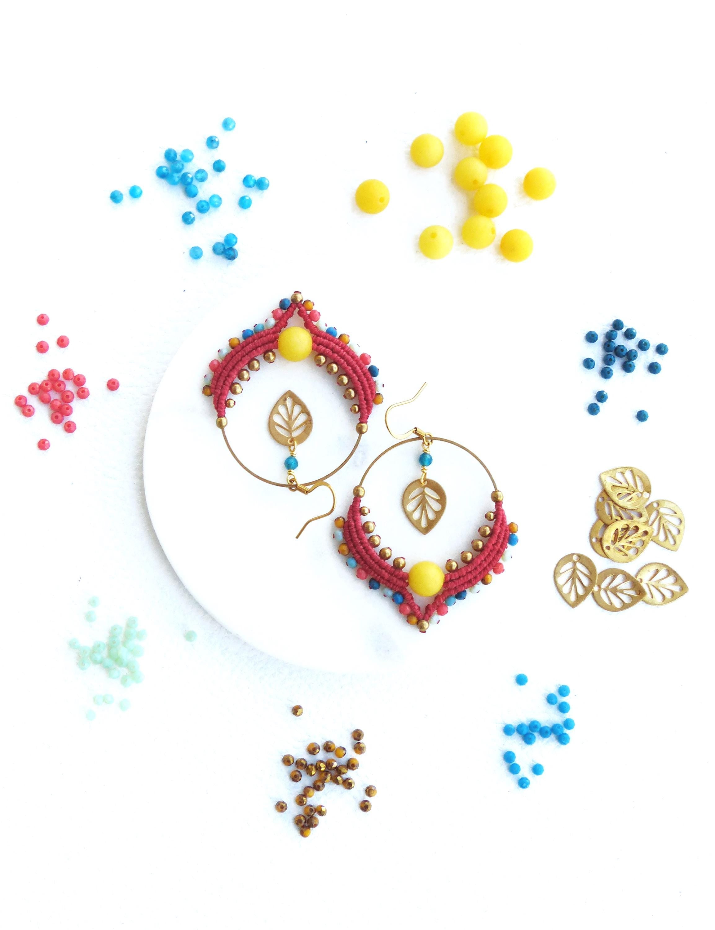 Peut inclure: Une paire de boucles d'oreilles faites à la main avec des perles rouges, bleues et jaunes, des accents de feuilles d'or et une perle jaune au centre. Les boucles d'oreilles sont sur une surface blanche avec des perles en vrac de différentes couleurs.