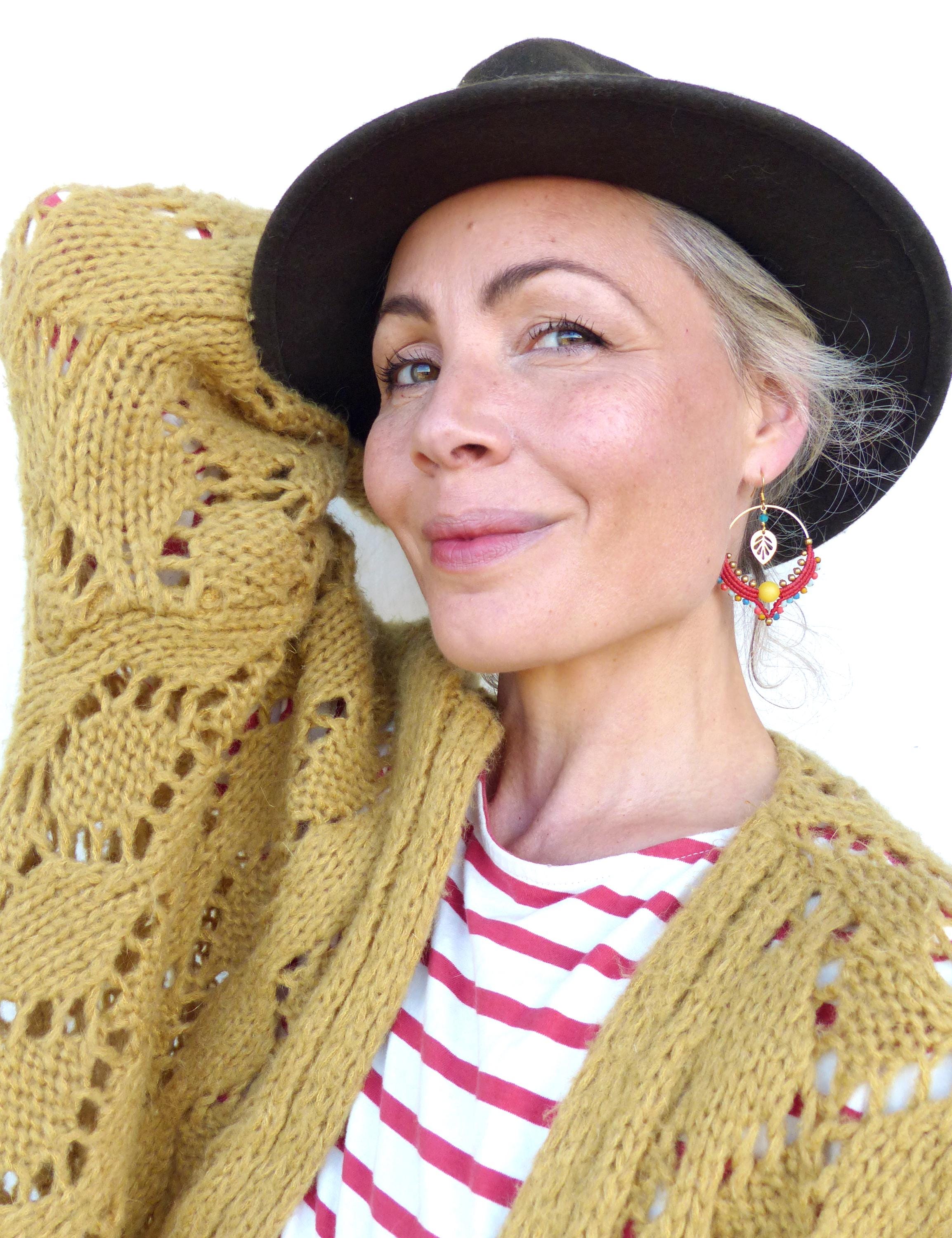 Peut inclure: Une femme portant un chapeau marron, un cardigan jaune et une chemise rayée. Elle porte des boucles d'oreilles en perles colorées avec un charme en forme de feuille.