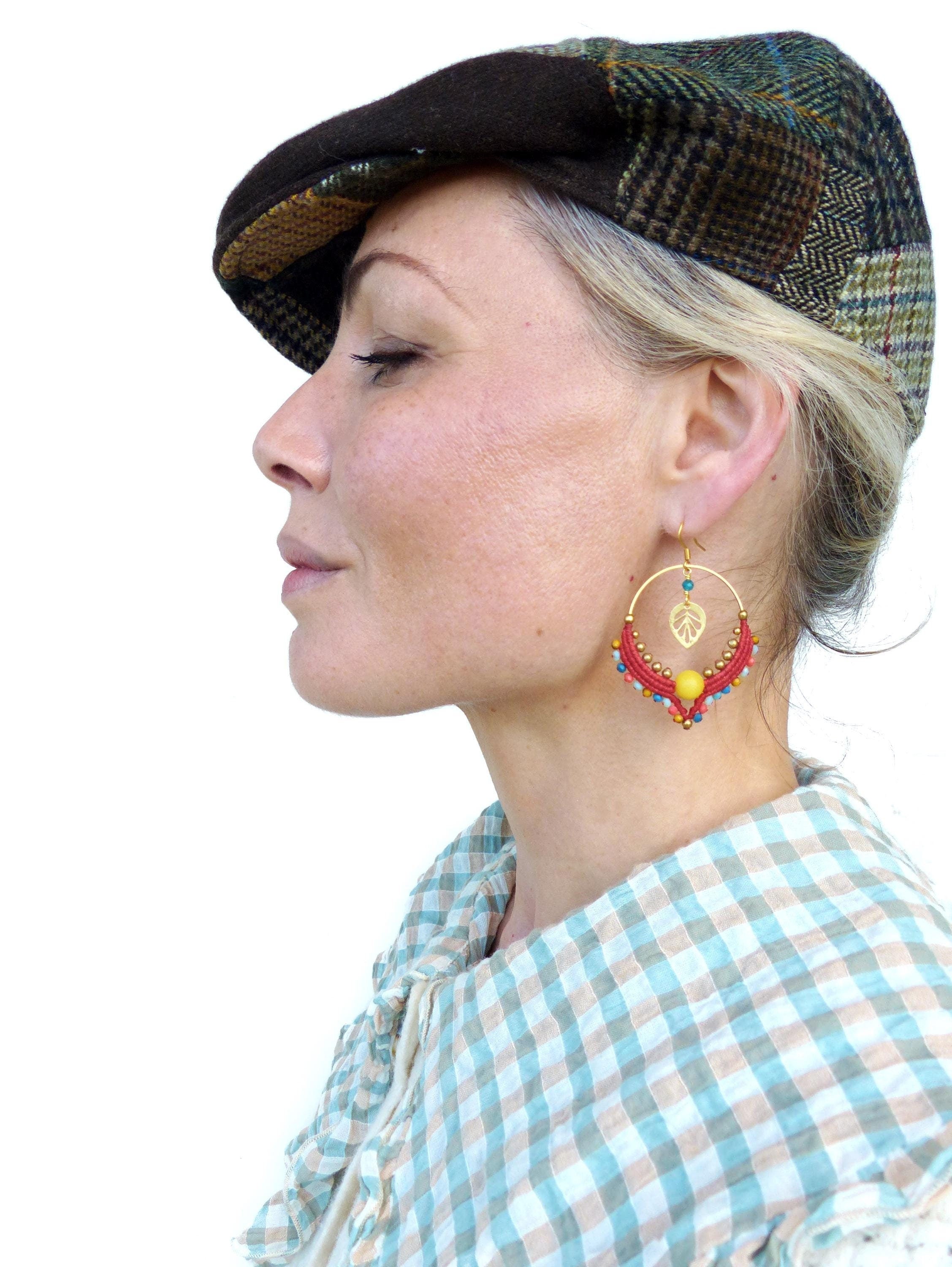 Peut inclure: Une femme portant une casquette de newsboy en tissu écossais brun et vert et une chemise à carreaux bleus et blancs. Elle porte des boucles d'oreilles créoles rouges et dorées avec une perle jaune et un pendentif en forme de feuille dorée.