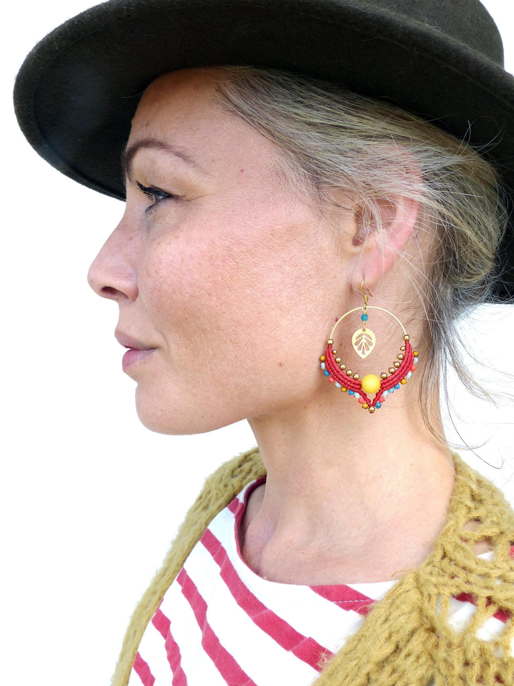 Peut inclure: Une femme portant une chemise à rayures rouges et blanches et un chapeau marron avec une paire de boucles d'oreilles créoles rouges et dorées avec un motif de feuille et des perles colorées.