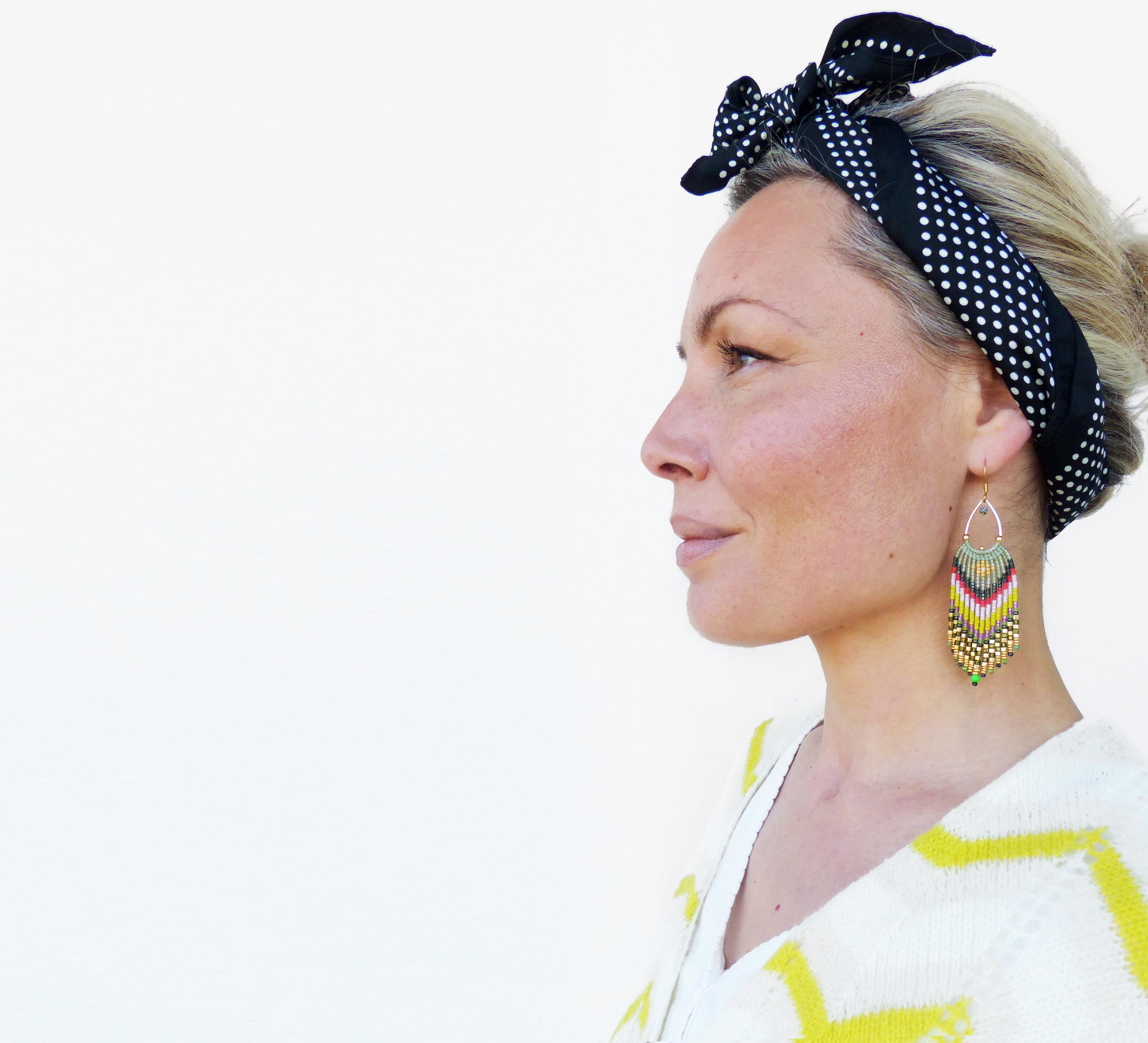 Peut inclure: Une femme aux cheveux blonds porte un bandeau à pois noirs et blancs et un pull blanc à rayures jaunes. Elle porte de longues boucles d'oreilles en perles avec des accents verts, rouges et dorés.