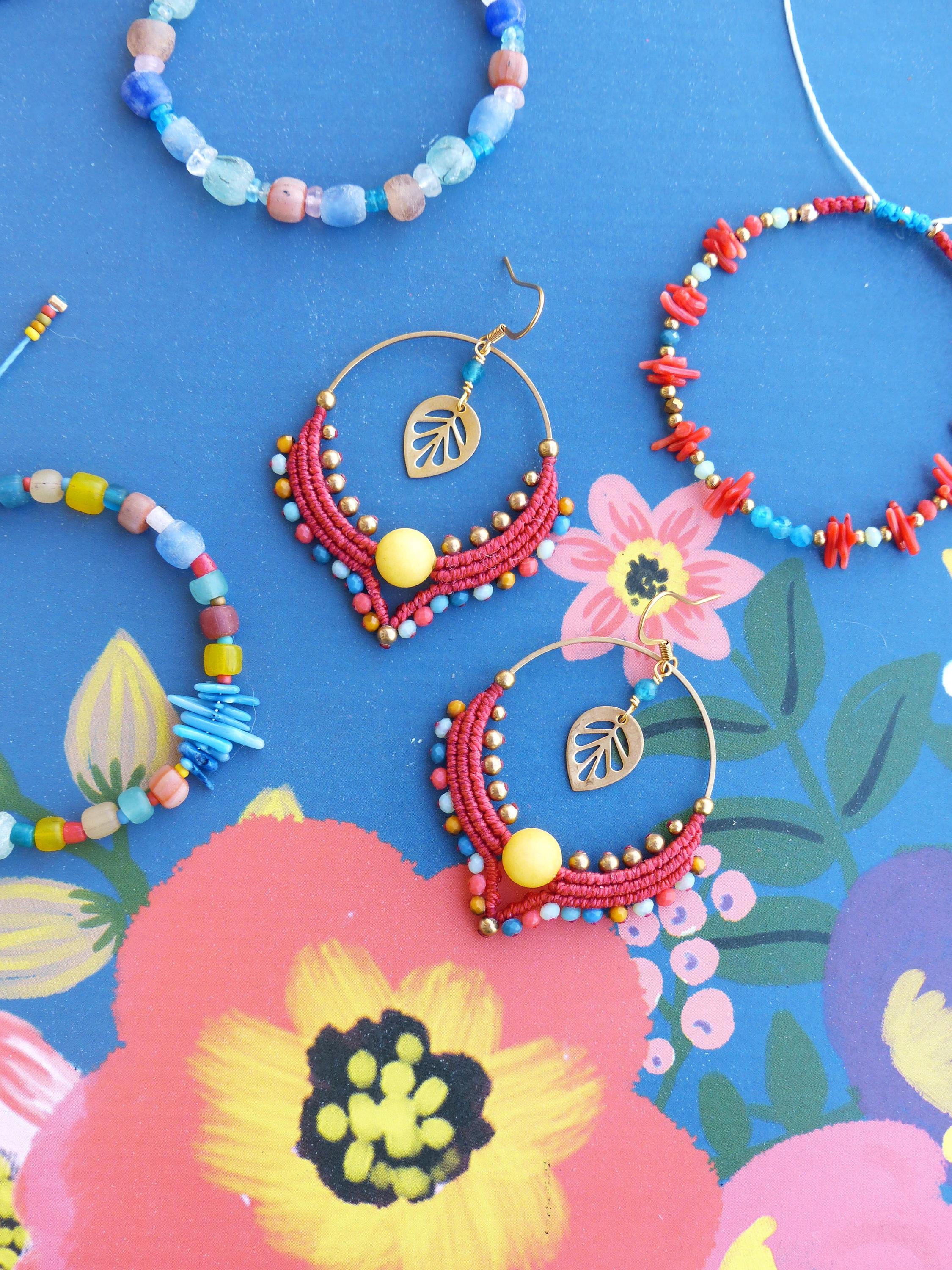 Peut inclure: Une paire de boucles d'oreilles créoles rouges et dorées avec une perle jaune et un pendentif en forme de feuille. Les boucles d'oreilles sont sur un fond bleu avec des motifs floraux.