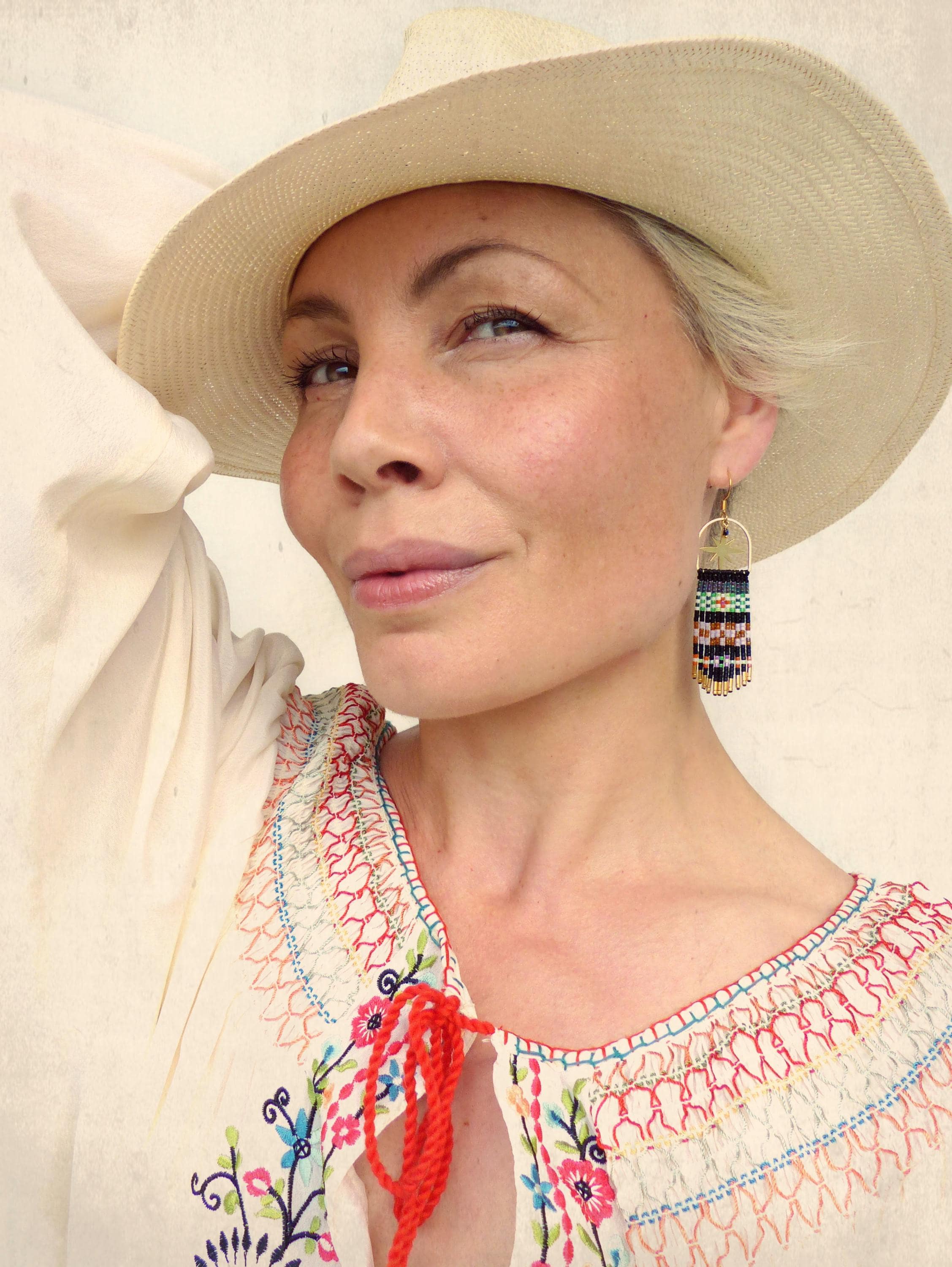 Peut inclure: Une femme portant une blouse blanche brodée avec un ruban rouge et un chapeau de paille à larges bords. Elle porte des boucles d'oreilles en perles avec des accents noirs, blancs, verts et dorés.