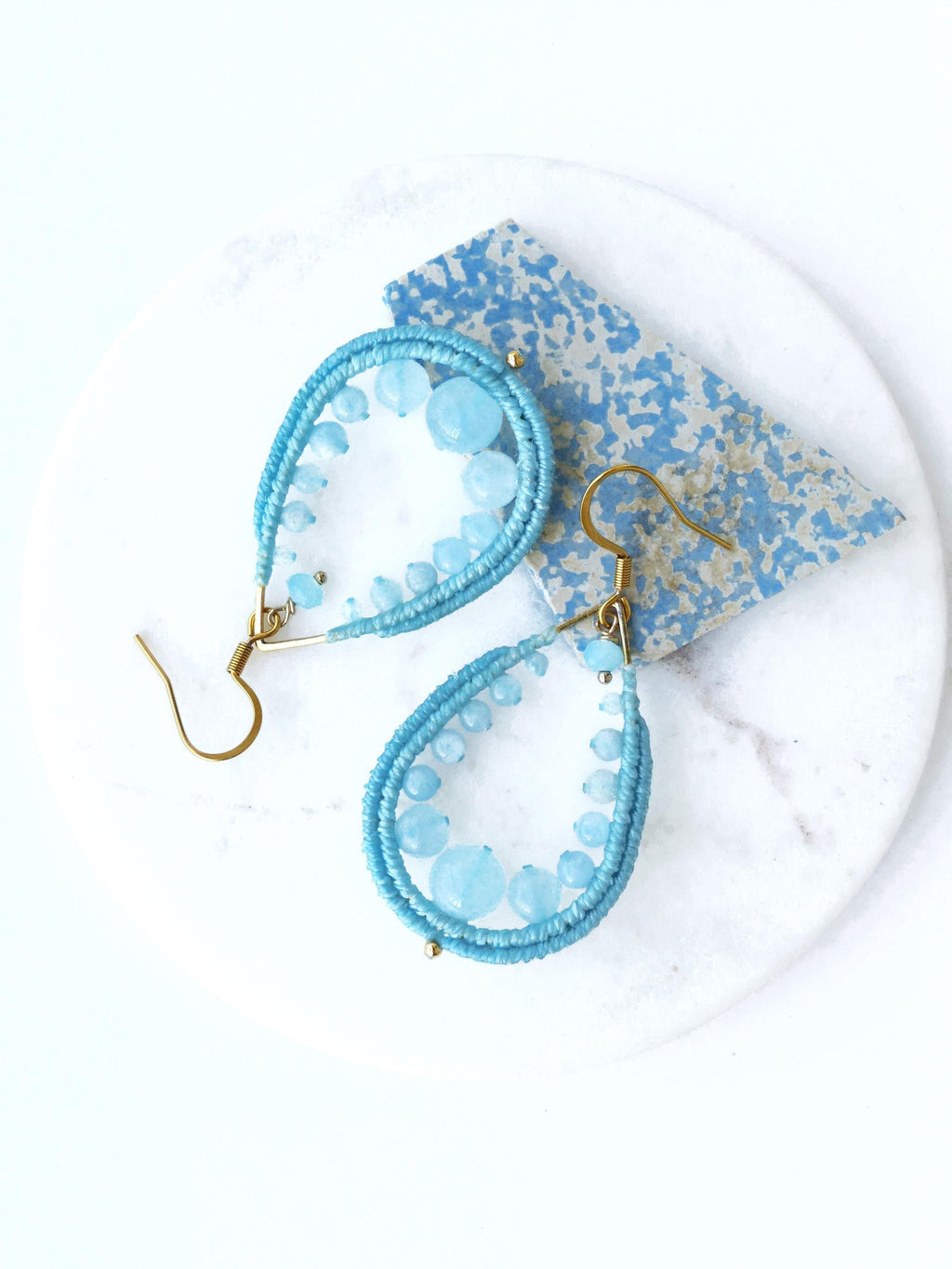 Peut inclure: Une paire de boucles d'oreilles en forme de goutte d'eau avec des perles bleues et du fil d'or. Les boucles d'oreilles sont sur une surface blanche.