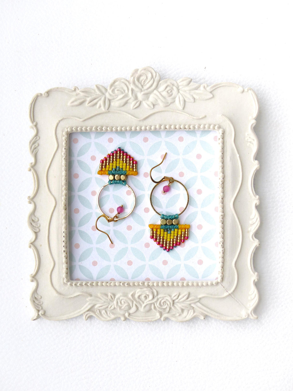 Peut inclure: Une paire de boucles d'oreilles faites à la main présentée dans un cadre blanc orné. Les boucles d'oreilles ont des cerceaux dorés avec des détails perlés colorés en rouge, jaune, bleu et rose. Le fond présente un motif de formes roses et bleues.