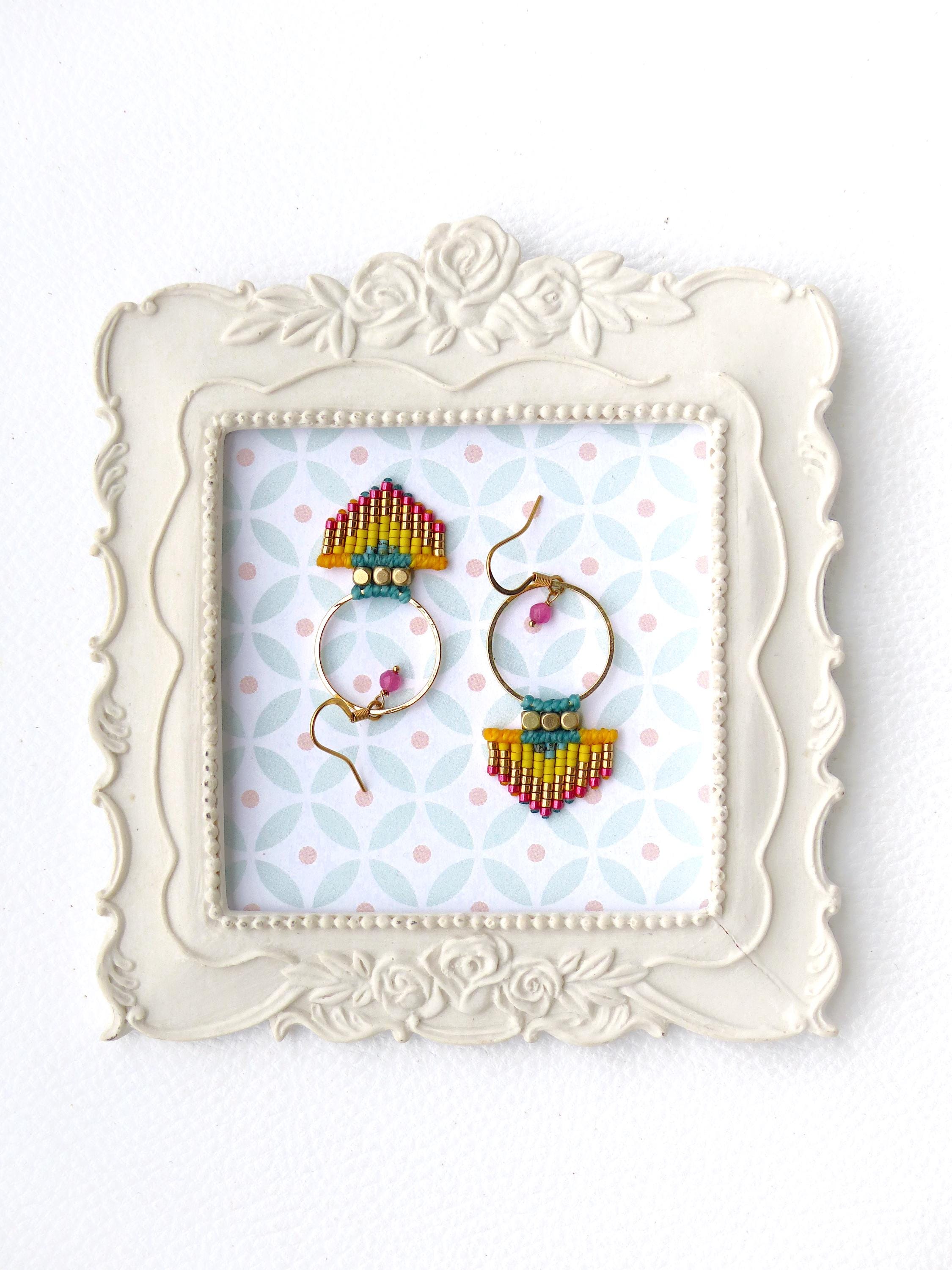 Peut inclure: Une paire de boucles d'oreilles faites à la main présentée dans un cadre blanc orné. Les boucles d'oreilles ont des cerceaux dorés avec des détails perlés colorés en rouge, jaune, bleu et rose. Le fond présente un motif de formes roses et bleues.