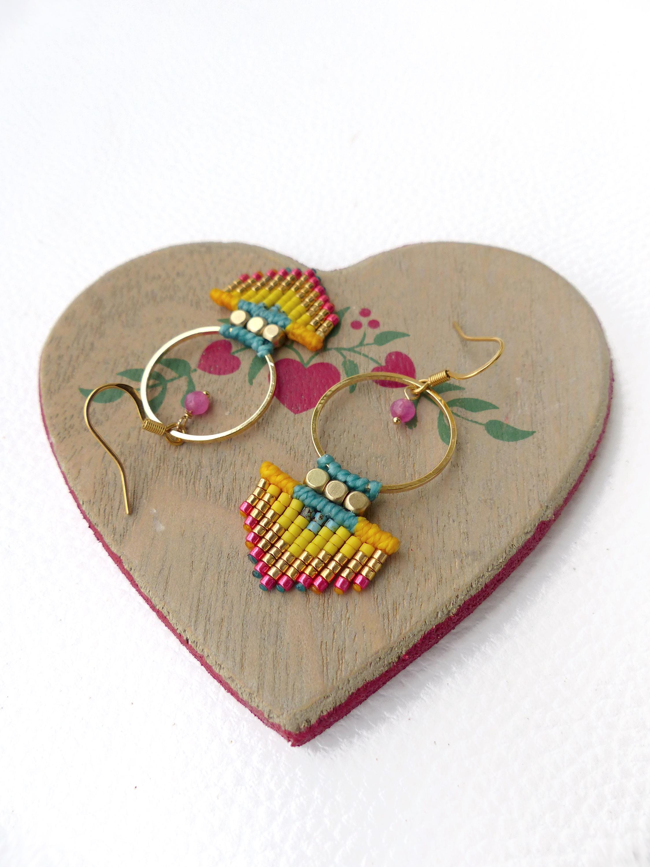 Peut inclure: Boucles d'oreilles faites à la main avec des cerceaux dorés et des détails perlés colorés. Les boucles d'oreilles présentent des perles jaunes, bleues et roses, avec une petite perle rose suspendue à chaque cercle. Présentées sur une surface en bois en forme de cœur.
