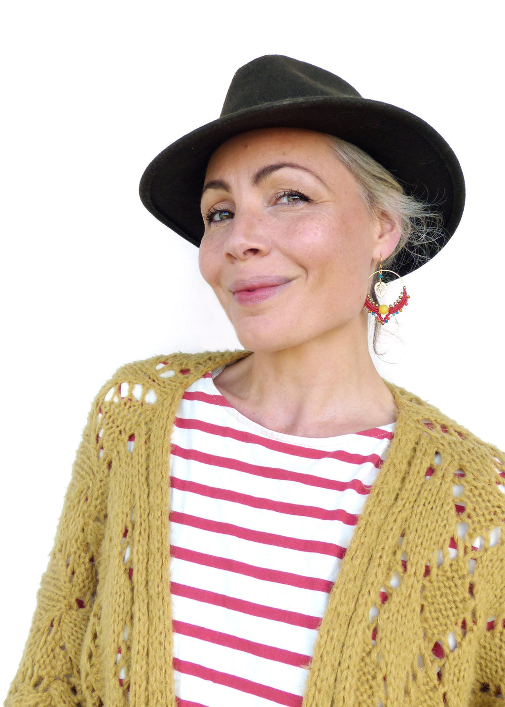 Peut inclure: Une femme portant un cardigan moutarde avec une chemise rayée rouge et blanche en dessous. Elle porte un chapeau fedora noir et des boucles d'oreilles en perles colorées.