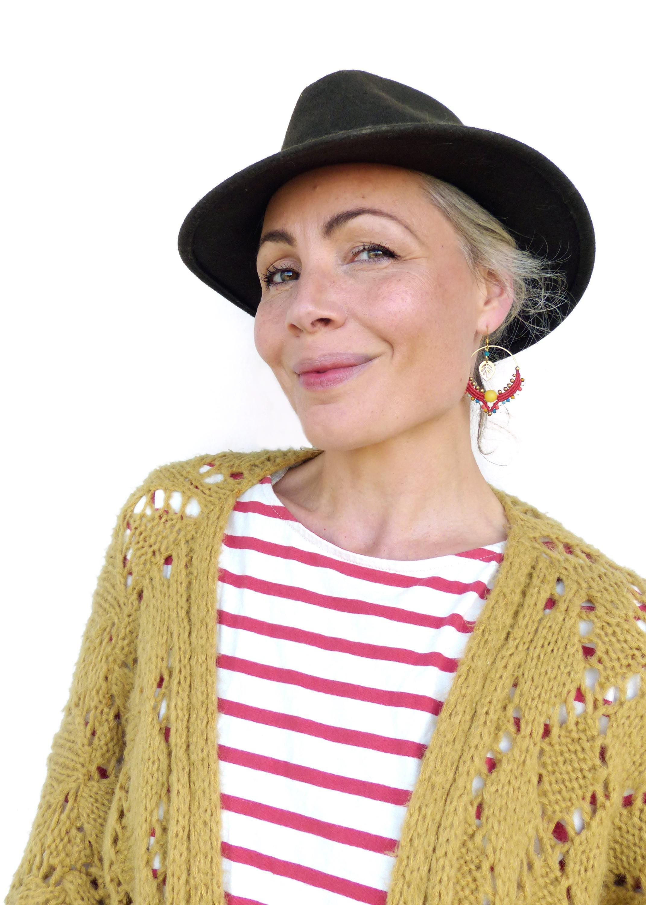 Peut inclure: Une femme portant un cardigan moutarde avec une chemise rayée rouge et blanche en dessous. Elle porte un chapeau fedora noir et des boucles d'oreilles en perles colorées.