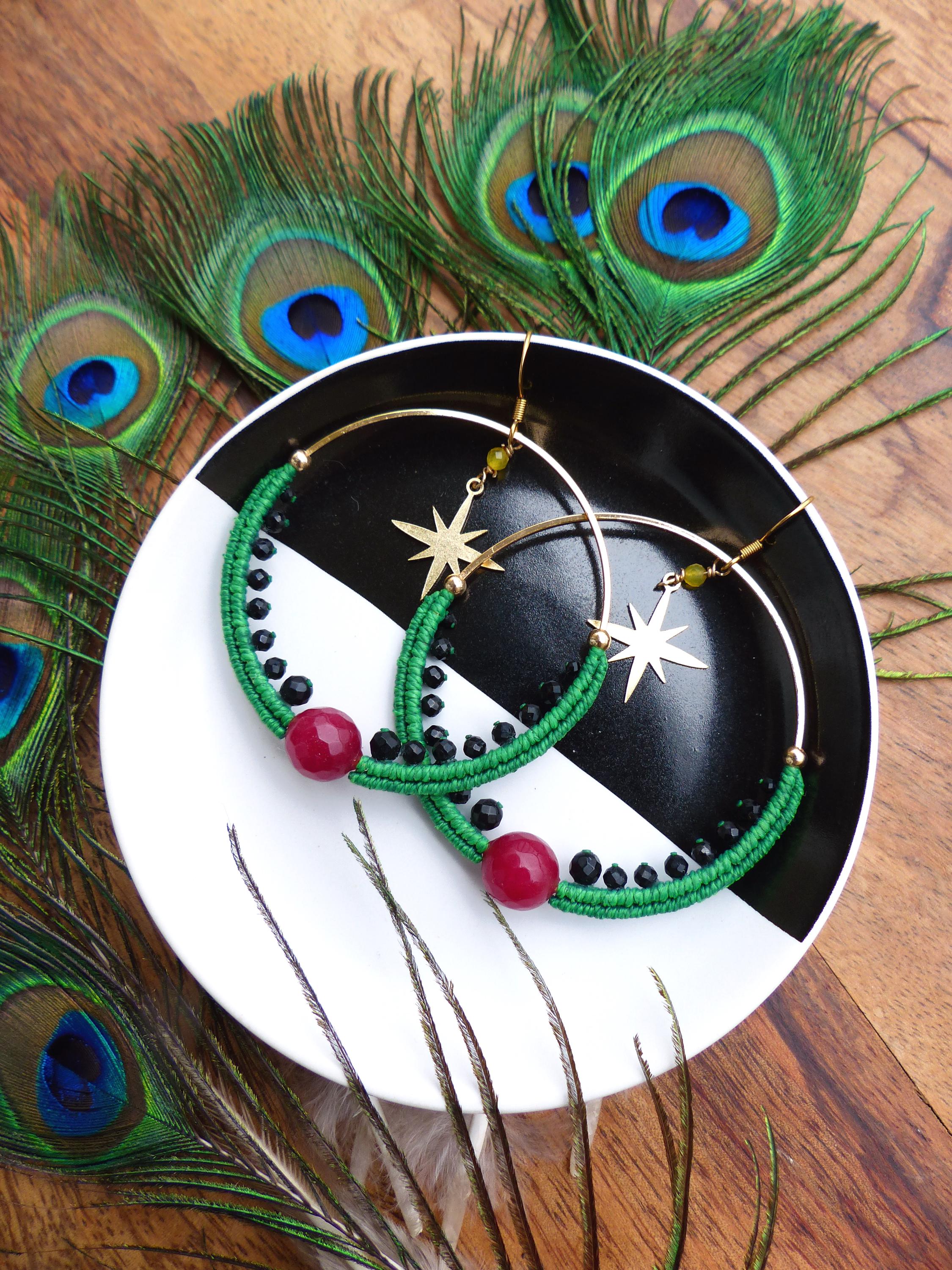 Peut inclure: Boucles d'oreilles créoles dorées avec macramé vert, perles rouges et noires, et breloques en forme d'étoile dorée. Les boucles d'oreilles sont posées sur une assiette noire et blanche, entourées de plumes de paon.