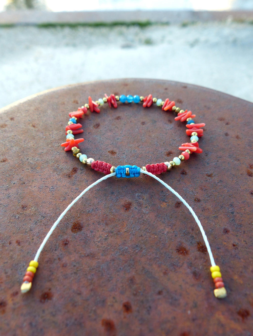 Peut inclure: Un bracelet en perles rouges, bleues et vertes avec un cordon blanc et des accents dorés. Le bracelet est noué et a un pompon à chaque extrémité.