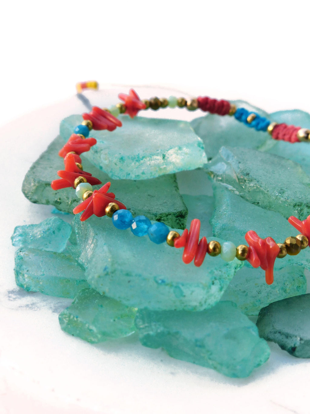 Peut inclure: Un bracelet en perles de corail rouge et turquoise avec des accents dorés, reposant sur un tas de verre de mer.