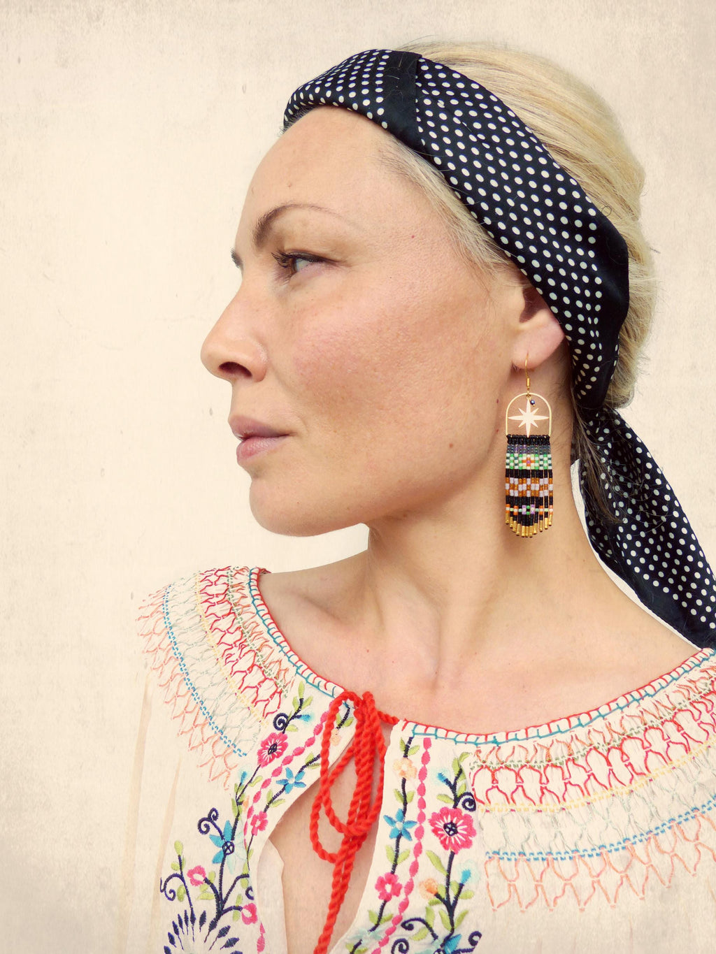 Peut inclure: Une femme portant un bandeau à pois noirs et blancs et une blouse blanche brodée avec des liens rouges. Elle porte de longues boucles d'oreilles en perles avec un charme d'étoile dorée.