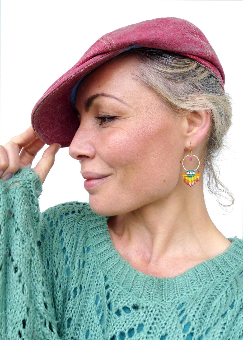 Peut inclure: Boucles d'oreilles créoles dorées avec un motif géométrique perlé jaune, vert et rouge. Les boucles d'oreilles ont une petite perle rose en haut. La femme porte une casquette marron et un pull vert d'eau.