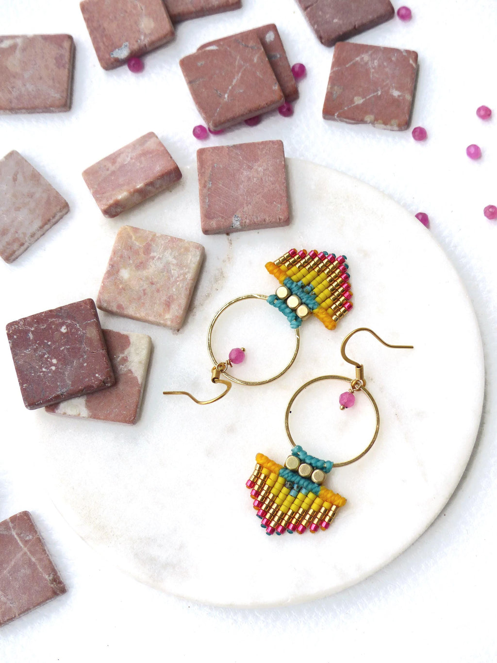 Peut inclure: Boucles d'oreilles macramé faites à la main avec des cerceaux dorés. Les boucles d'oreilles présentent un motif en forme d'éventail avec des perles jaunes, bleues et roses. De petites perles roses et des carreaux de pierre carrés sont dispersés sur une surface blanche.