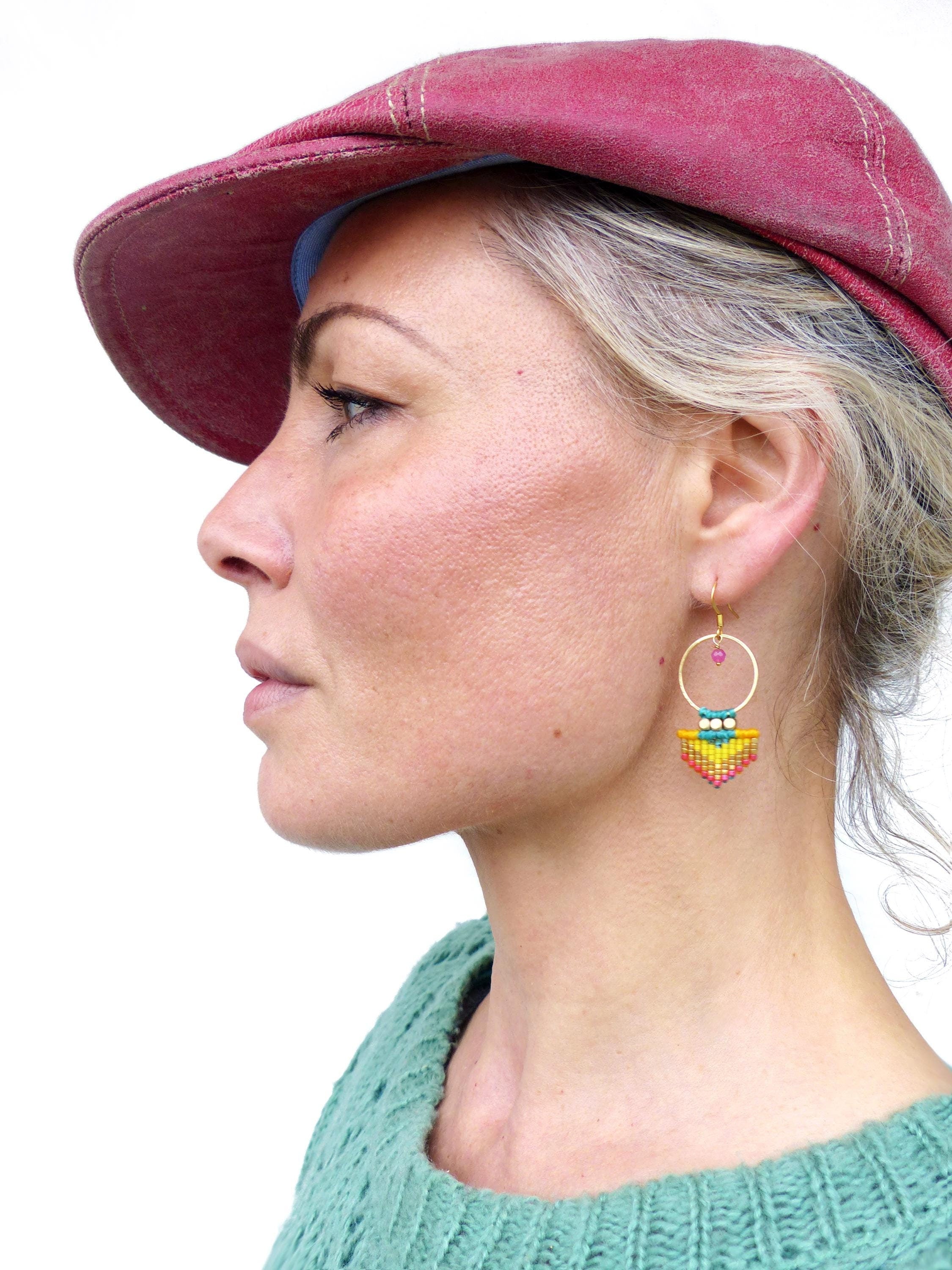 Peut inclure: Une femme porte une casquette gavroche bordeaux et un pull vert d'eau. Des boucles d'oreilles créoles dorées avec un motif de perles jaunes, oranges, roses et bleues pendent de son oreille. Une petite perle rose est au sommet de la boucle.