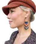 Peut inclure: Une femme porte une casquette de style gavroche rouille et une veste en tweed. L'accent est mis sur une paire de boucles d'oreilles à franges perlées avec des cerceaux dorés. Les boucles d'oreilles présentent un motif de perles bleues, oranges, vertes et blanches.