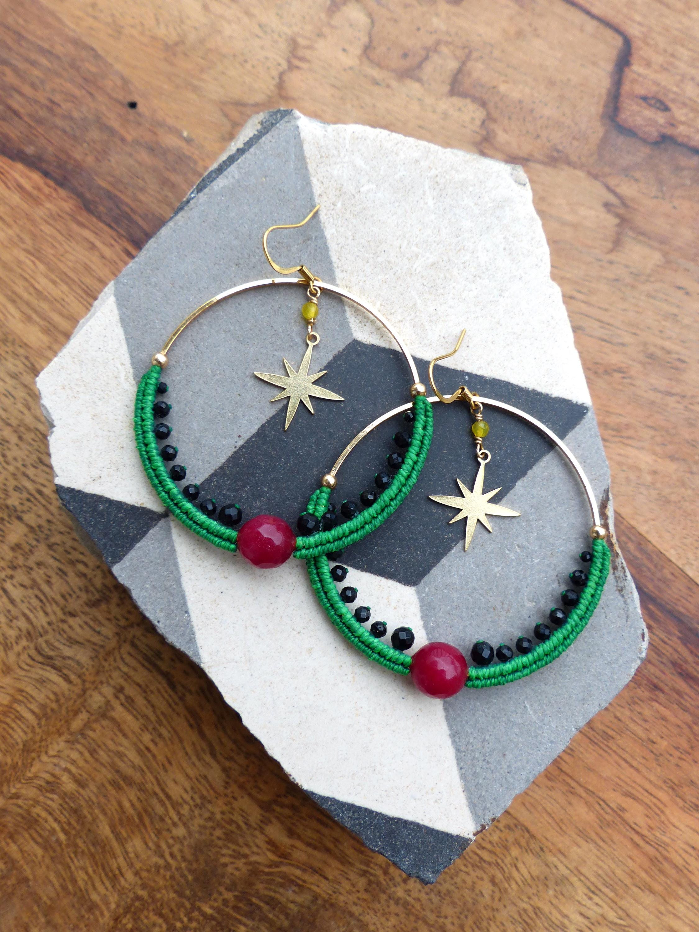 Peut inclure: Paire de boucles d'oreilles créoles dorées avec des détails en macramé vert, des perles noires et des accents de pierres rouges. Chaque boucle d'oreille comporte un charm en forme d'étoile dorée et une petite perle jaune. Présentées sur une dalle grise et blanche.