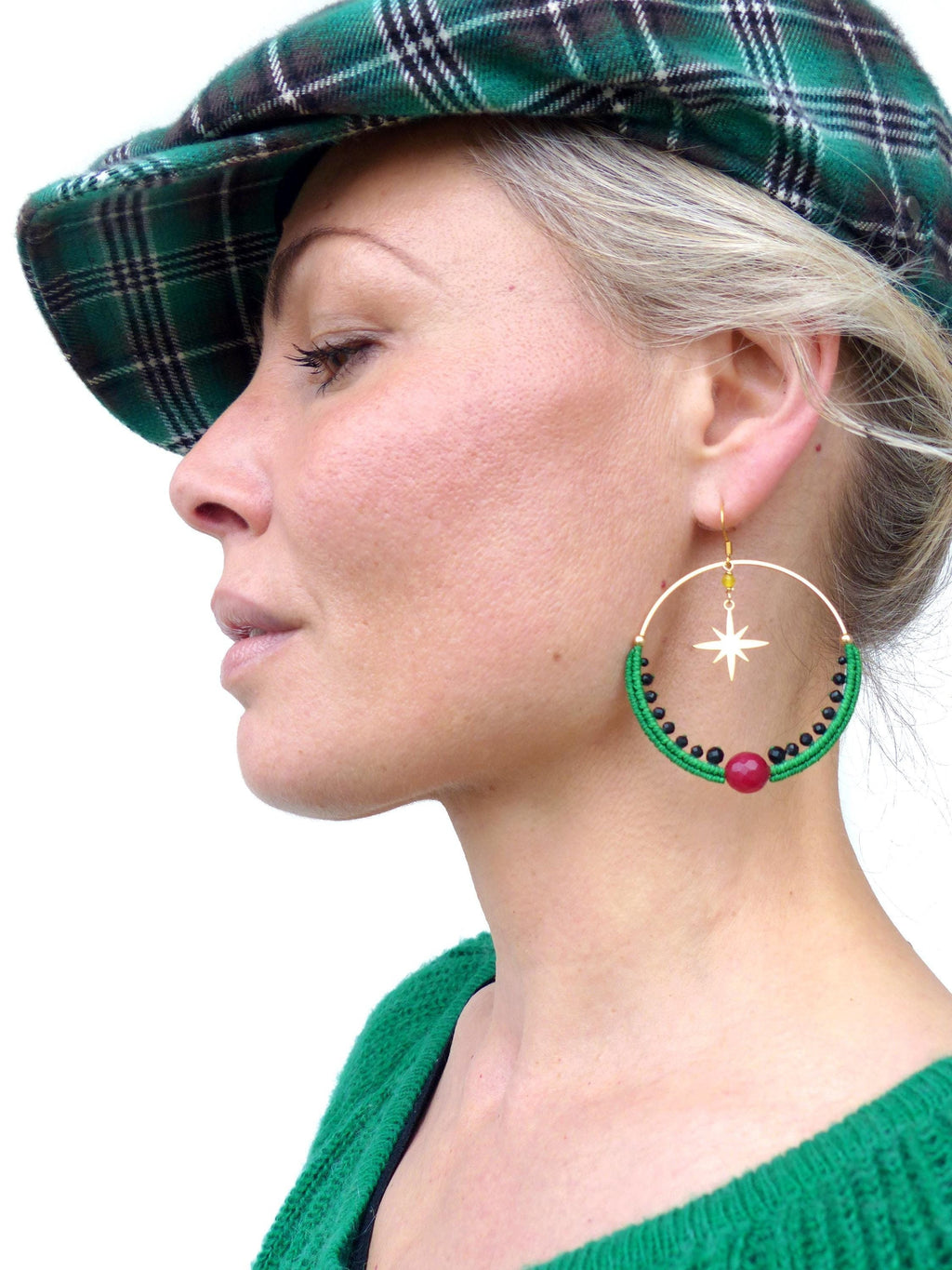 Peut inclure: Boucles d'oreilles créoles dorées avec un motif d'étoile, ornées de macramé vert, de perles noires et d'une pierre rouge. Elles sont portées avec un pull vert et un béret vert à carreaux.