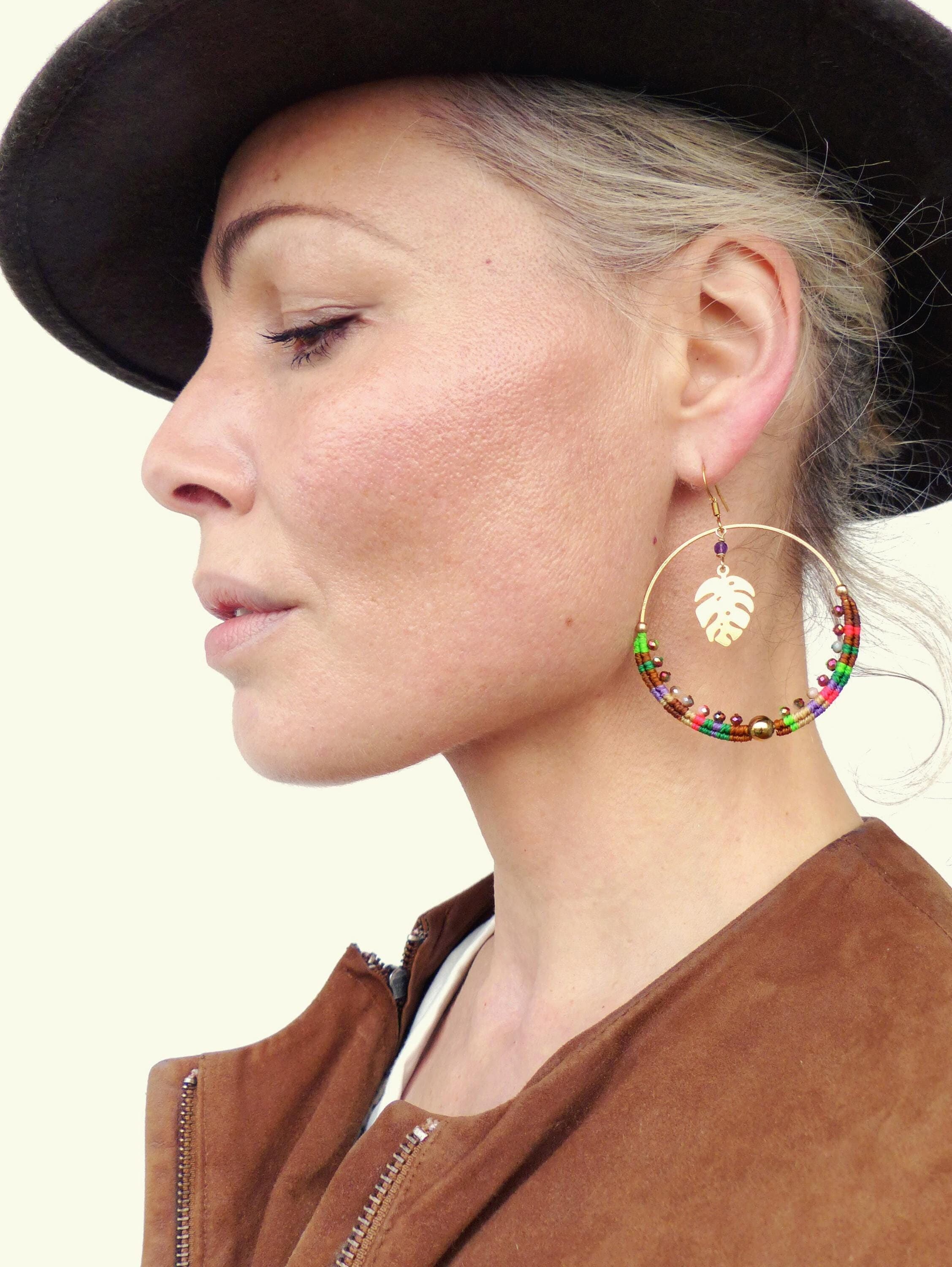 Peut inclure: Une femme portant un chapeau et une veste marrons avec de grandes boucles d'oreilles en forme de cerceau ornées d'un pendentif en forme de feuille d'or et de perles colorées.