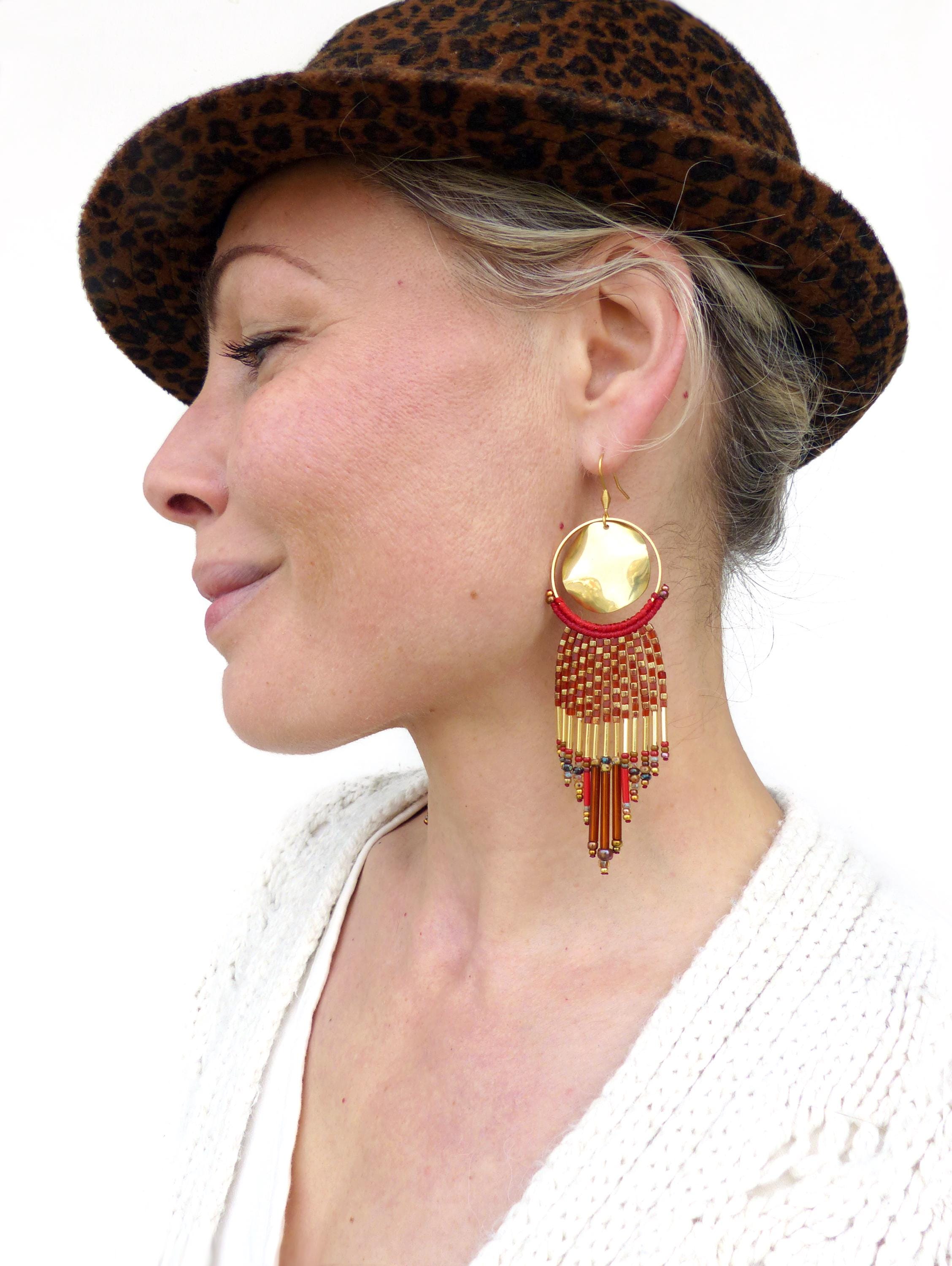 Peut inclure: Une paire de longues boucles d'oreilles en perles dorées et rouges avec un grand pendentif rond en or. Les boucles d'oreilles sont portées par une personne avec un chapeau léopard brun et noir.