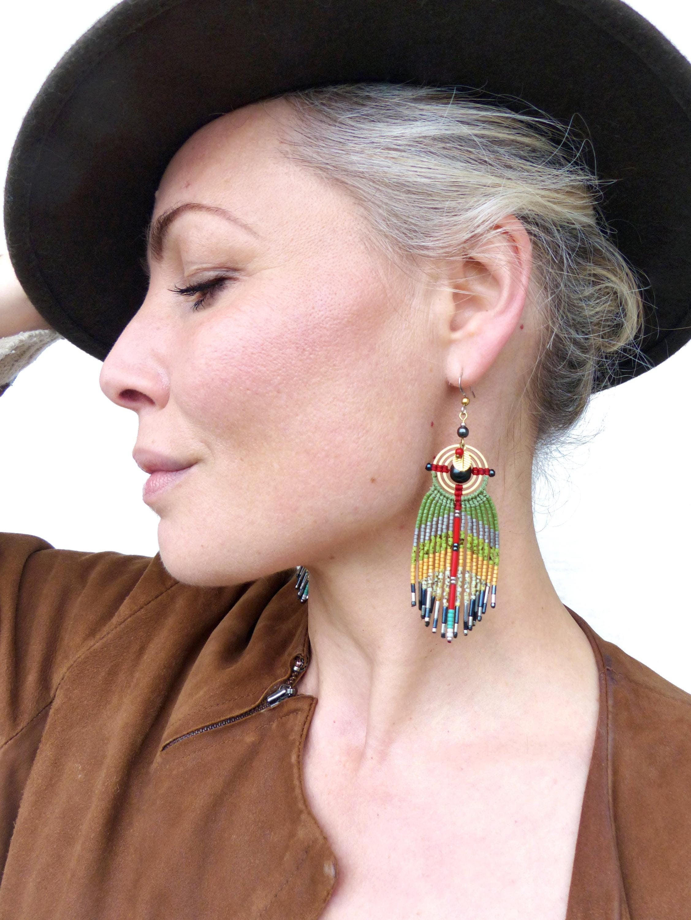 Peut inclure: Une femme portant une veste en daim marron et un chapeau noir avec de longs cheveux gris. Elle porte de longues boucles d'oreilles en perles colorées avec un motif géométrique or et noir.