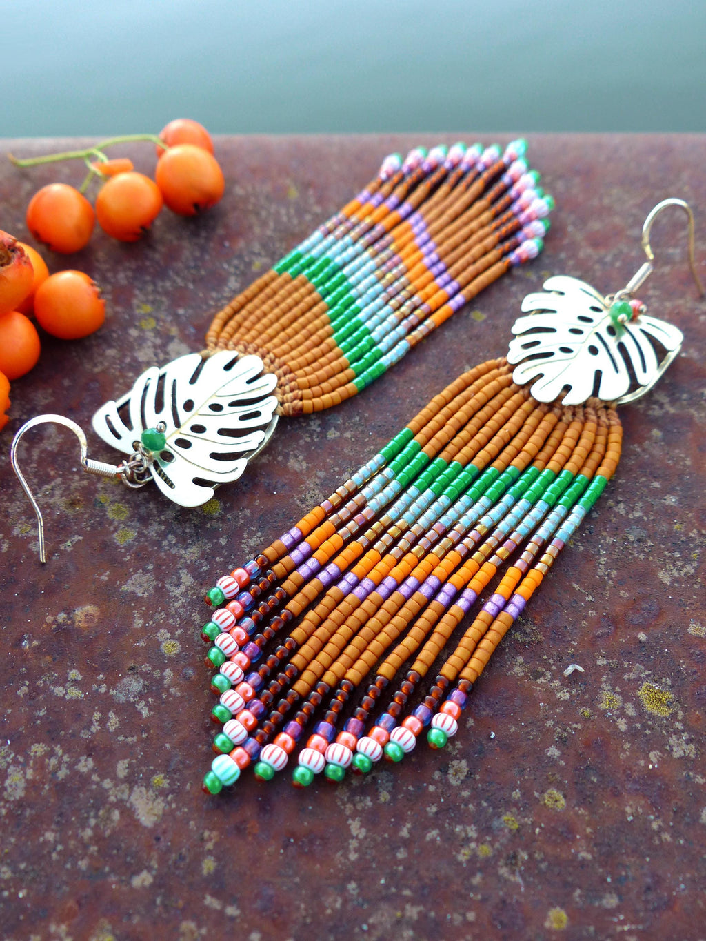 Peut inclure: Une paire de boucles d'oreilles faites à la main avec un motif de feuille tropicale. Les boucles d'oreilles présentent un dessus en forme de feuille de couleur argentée et une longue frange de perles marron avec des accents verts, oranges, violets et roses.
