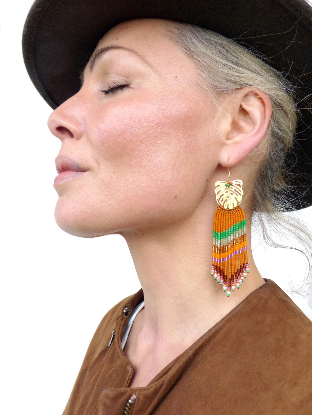 Peut inclure: Boucles d'oreilles dorées avec un motif de feuille et des franges de perles colorées dans les tons orange, vert et rose. Les boucles d'oreilles sont suspendues à l'oreille d'une personne. La personne porte une veste marron et un chapeau foncé.