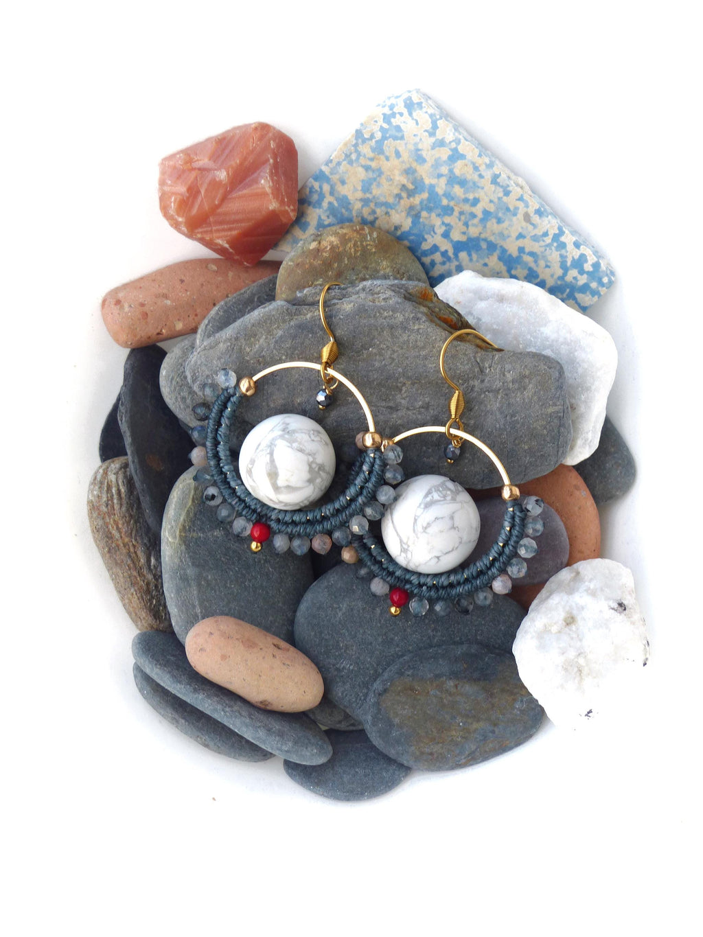 Peut inclure: Boucles d'oreilles créoles faites à la main avec une monture en métal doré. Les boucles d'oreilles présentent une grosse perle blanche effet marbre au centre, entourée d'un motif macramé bleu foncé et de petites perles rouges et grises. Elles sont posées sur des pierres grises et blanches.