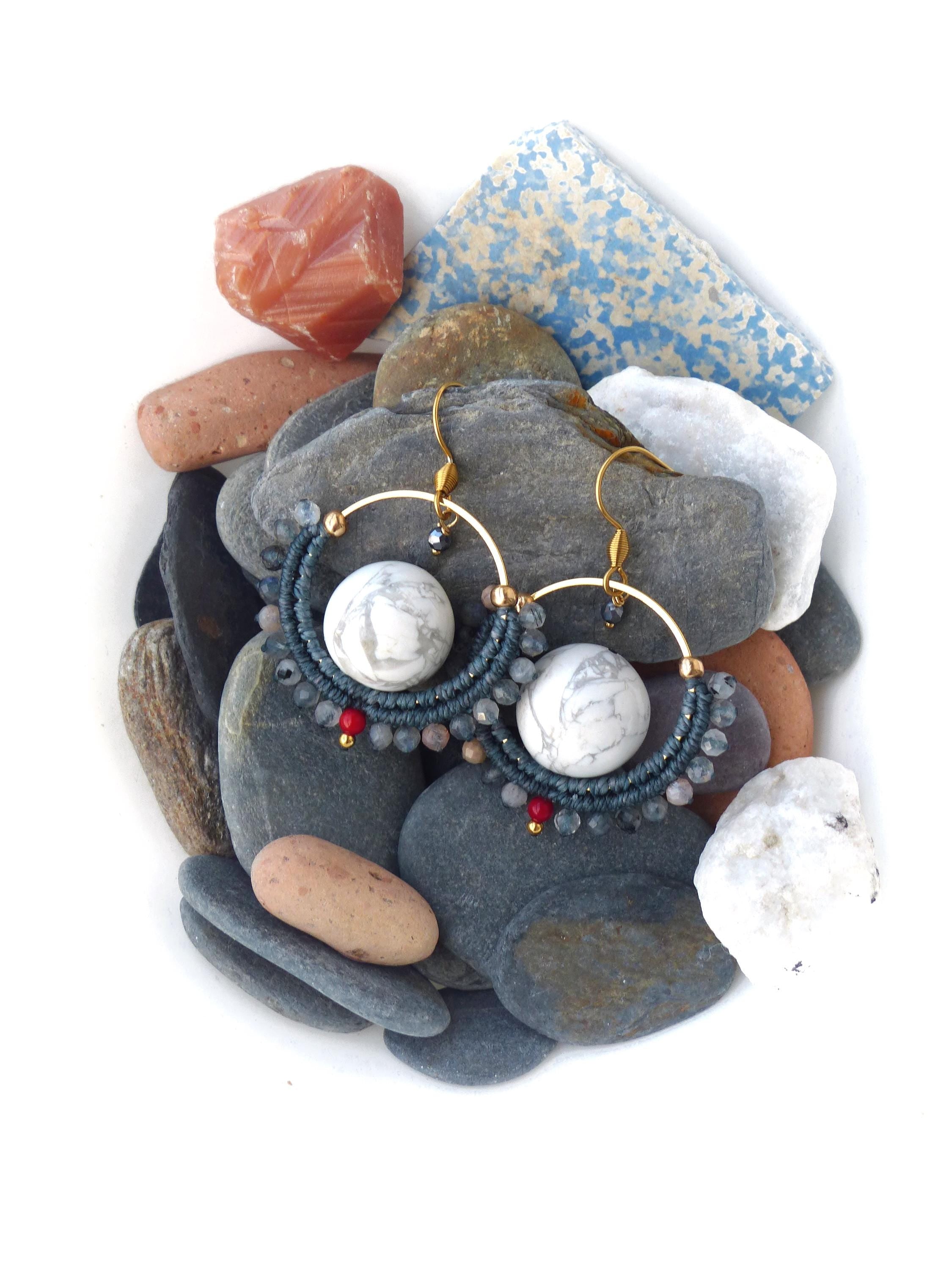 Peut inclure: Boucles d'oreilles créoles faites à la main avec une monture en métal doré. Les boucles d'oreilles présentent une grosse perle blanche effet marbre au centre, entourée d'un motif macramé bleu foncé et de petites perles rouges et grises. Elles sont posées sur des pierres grises et blanches.