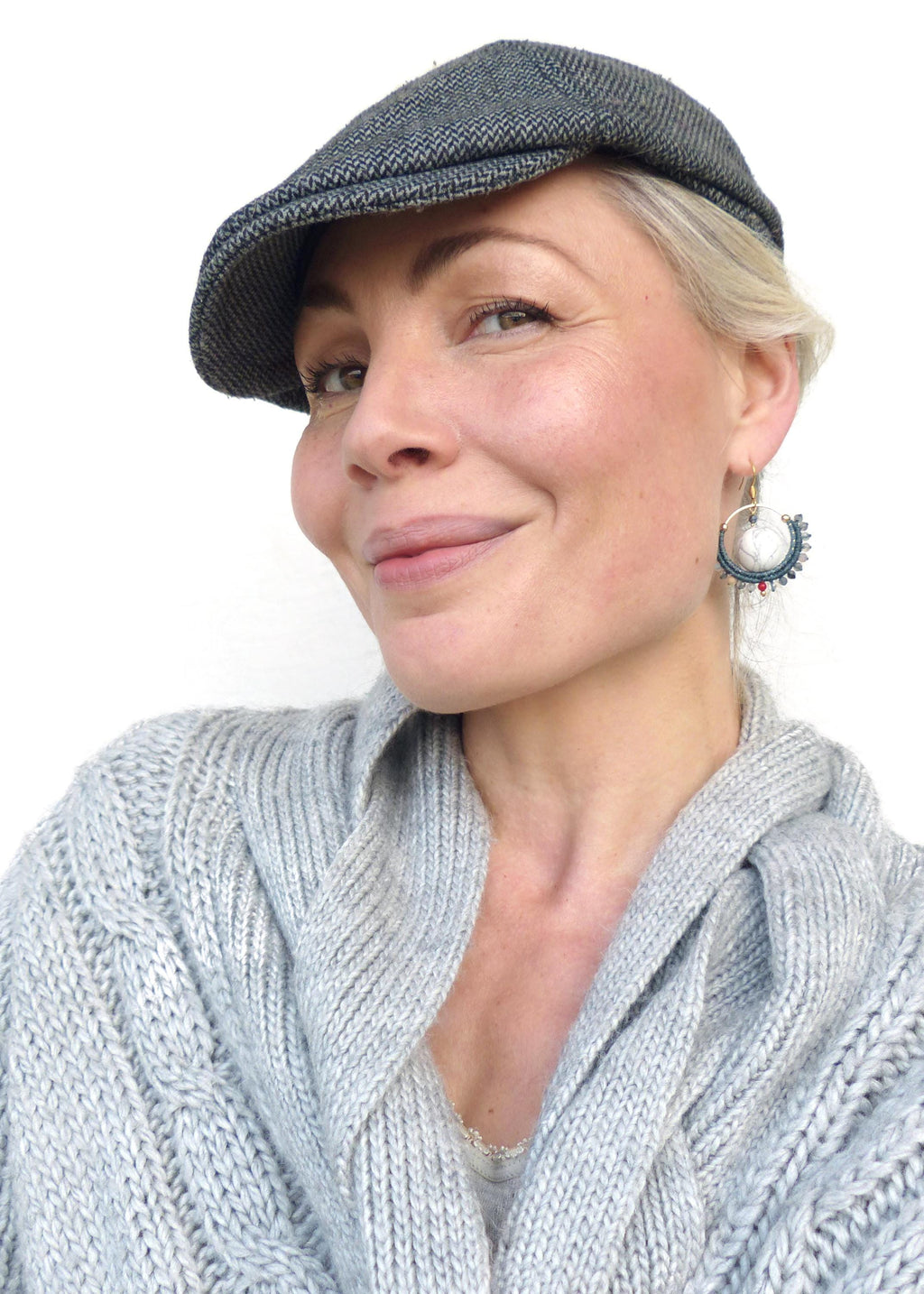 Peut inclure: Une femme portant une casquette gavroche en tweed à chevrons gris et un pull en tricot torsadé gris clair. Elle porte également des boucles d'oreilles créoles avec des accents blancs, bleus et rouges.