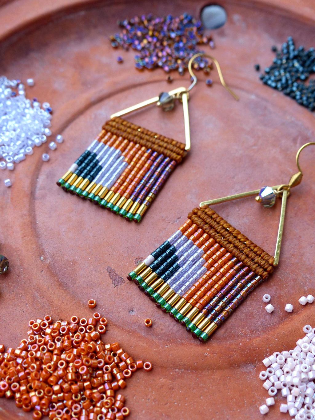 Peut inclure: Une paire de boucles d'oreilles perlées faites à la main avec un motif géométrique. Les boucles d'oreilles présentent une section supérieure marron et une frange colorée de perles dans les tons orange, violet, vert et or. Elles sont suspendues à des crochets dorés.