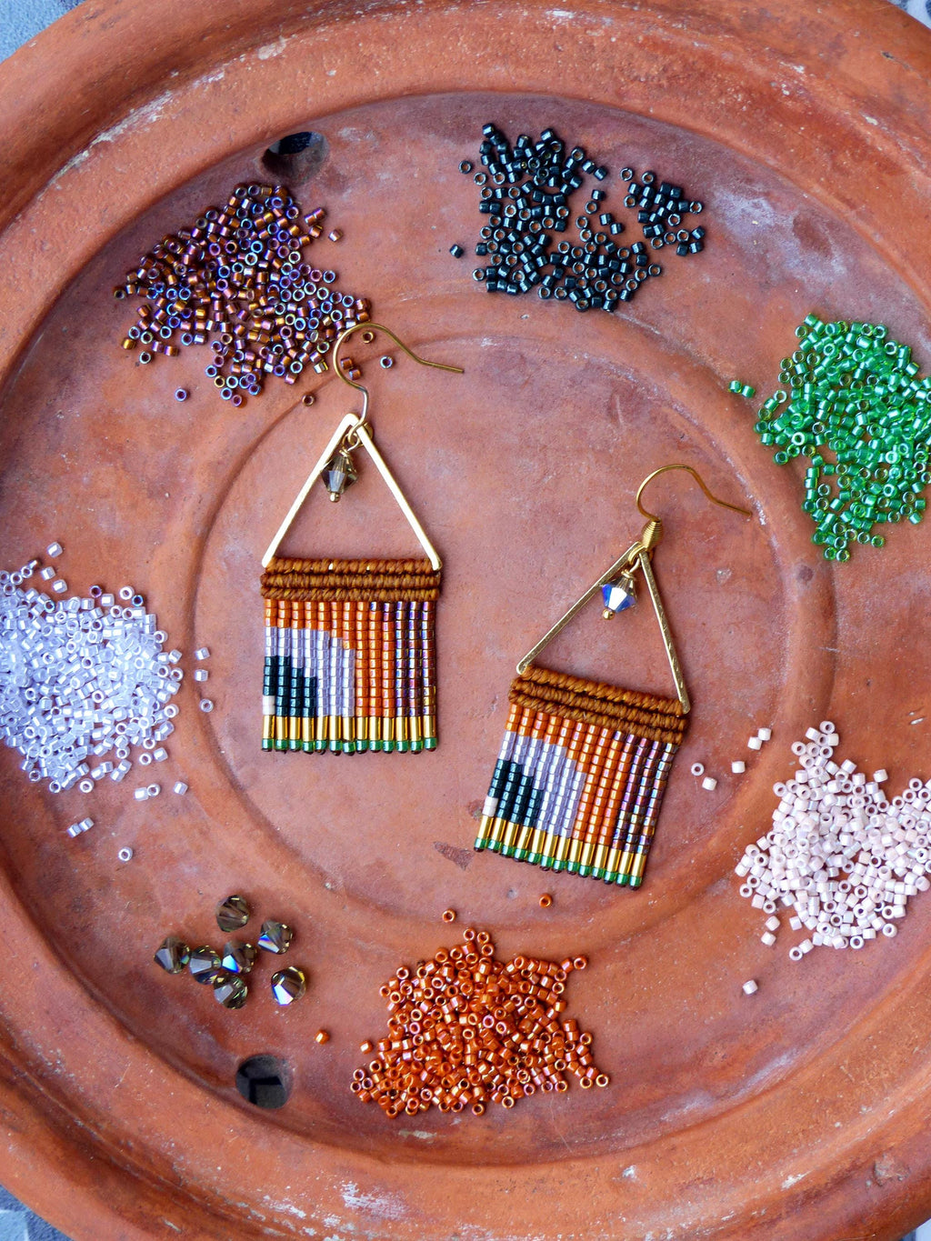 Peut inclure: Boucles d'oreilles faites à la main avec un motif géométrique. Les boucles d'oreilles présentent un cadre triangulaire doré et sont ornées de rangées de perles orange, marron, vertes et violettes. Divers tas de perles colorées entourent les boucles d'oreilles sur une surface en terre cuite.