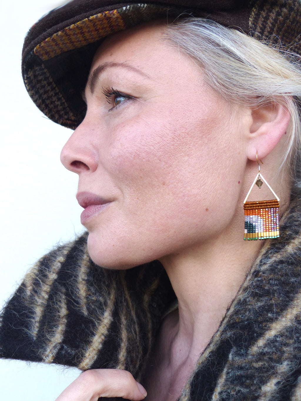 Peut inclure: Profil d'une personne portant une casquette en tweed marron et un manteau à motifs. La personne porte des boucles d'oreilles triangulaires dorées avec un motif de perles colorées. Les boucles d'oreilles présentent des perles orange, vertes et bleues.