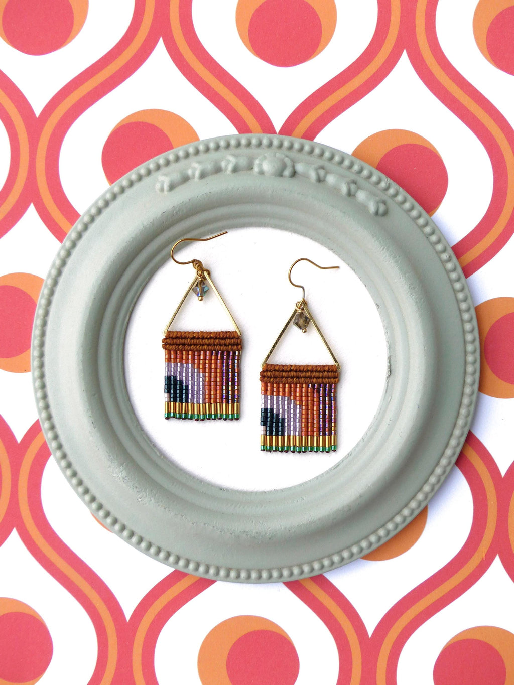 Peut inclure: Une paire de boucles d'oreilles perlées faites à la main. Les boucles d'oreilles présentent un cadre triangulaire doré avec une frange de perles colorées dans les tons marron, orange, violet, vert et noir. Elles sont présentées dans un cadre rond bleu clair.