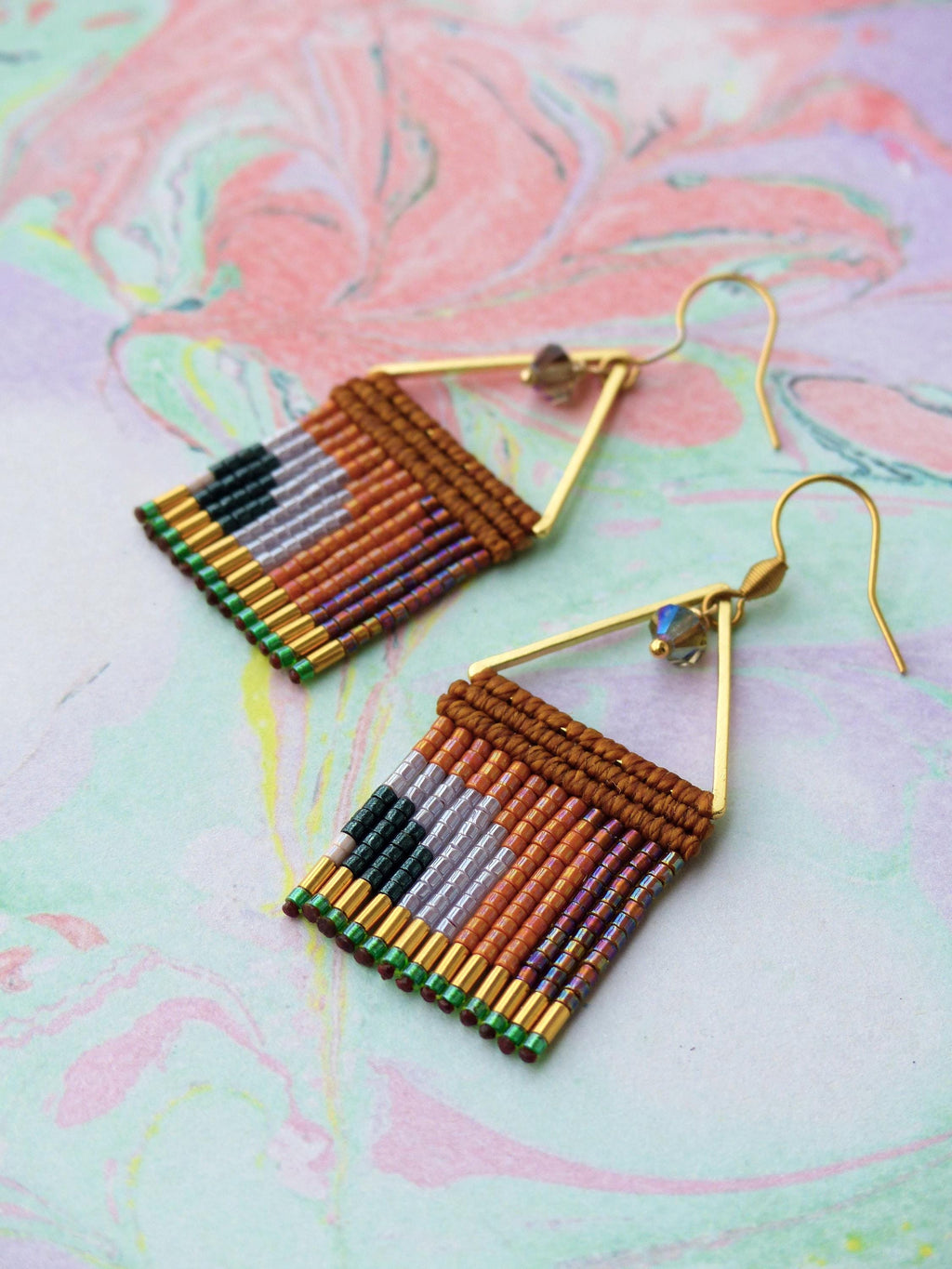 Peut inclure: Une paire de boucles d'oreilles faites à la main avec un motif géométrique. Les boucles d'oreilles présentent un cadre triangulaire doré et un motif tissé de perles marron, orange, violet, gris, vert et noir. Elles ont une fermeture à crochet et une petite perle de cristal.