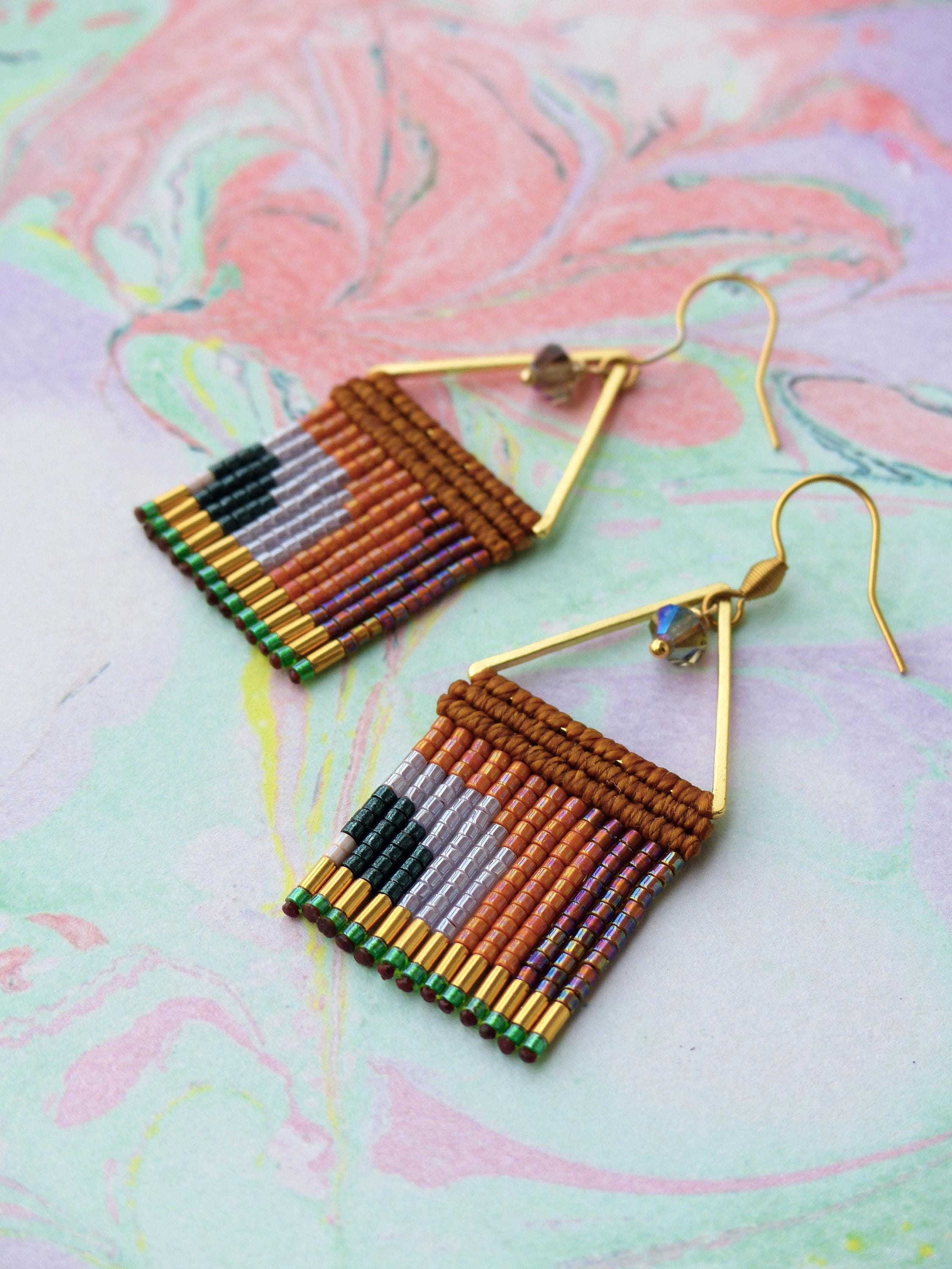 Peut inclure: Une paire de boucles d'oreilles faites à la main avec un motif géométrique. Les boucles d'oreilles présentent un cadre triangulaire doré et un motif tissé de perles marron, orange, violet, gris, vert et noir. Elles ont une fermeture à crochet et une petite perle de cristal.