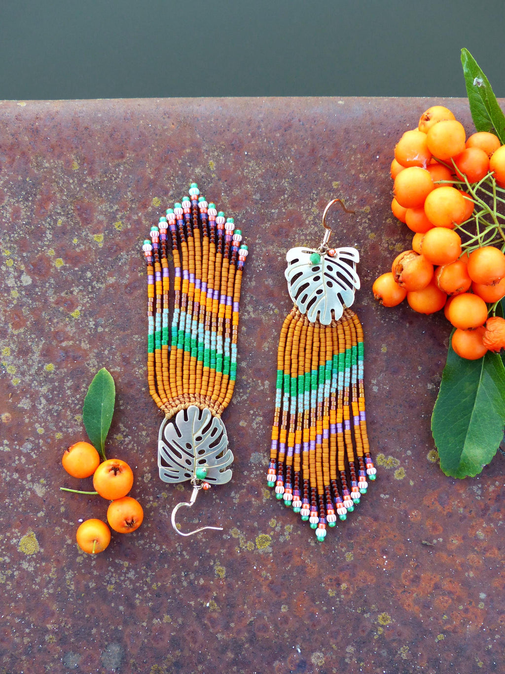 Peut inclure: Boucles d'oreilles faites à la main avec un motif de feuille tropicale. Les boucles d'oreilles présentent des perles orange, vertes, violettes et marron. La feuille est de couleur argentée. Les boucles d'oreilles sont présentées sur une surface métallique rouillée avec des baies oranges.