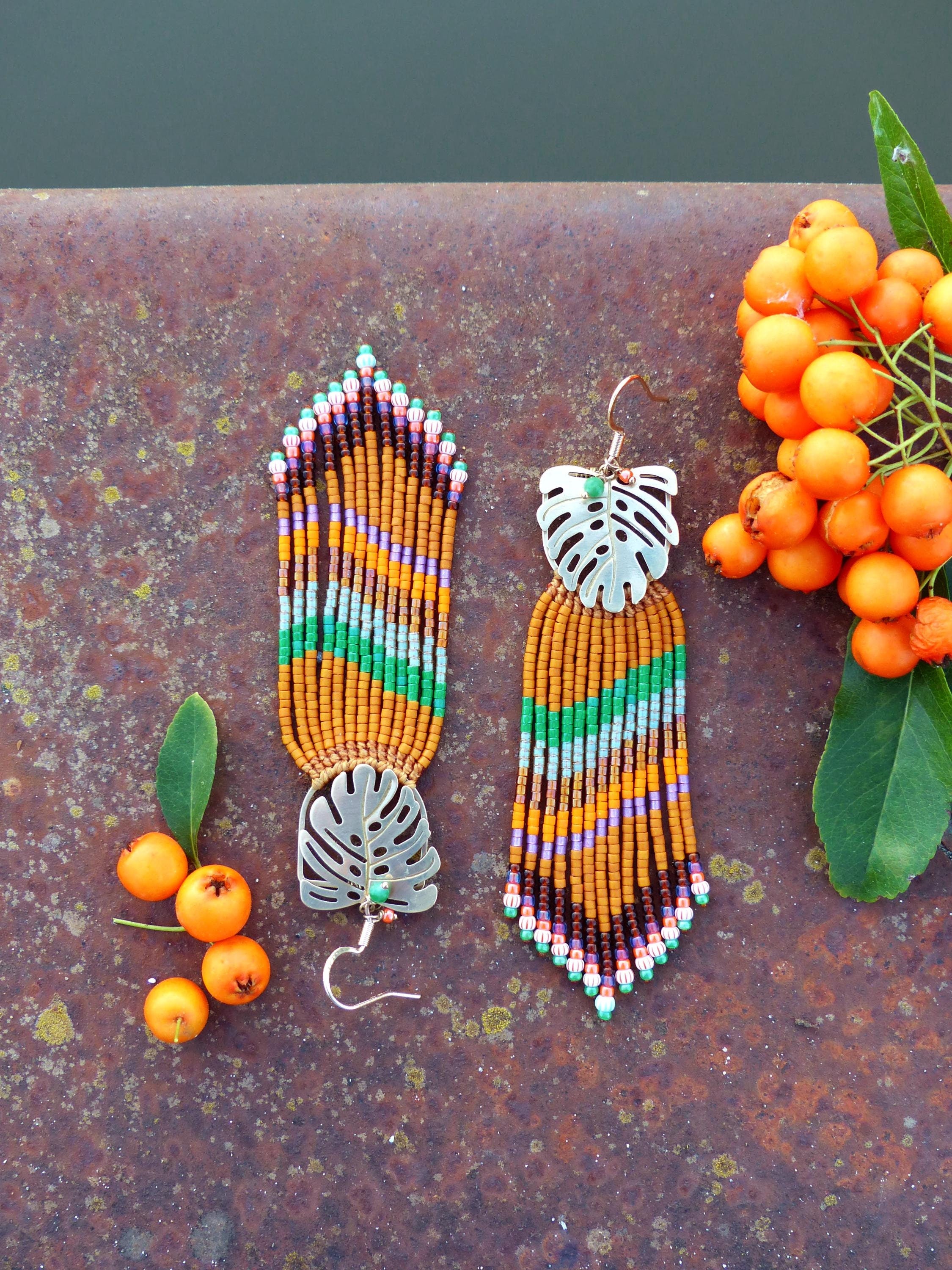 Peut inclure: Boucles d'oreilles faites à la main avec un motif de feuille tropicale. Les boucles d'oreilles présentent des perles orange, vertes, violettes et marron. La feuille est de couleur argentée. Les boucles d'oreilles sont présentées sur une surface métallique rouillée avec des baies oranges.