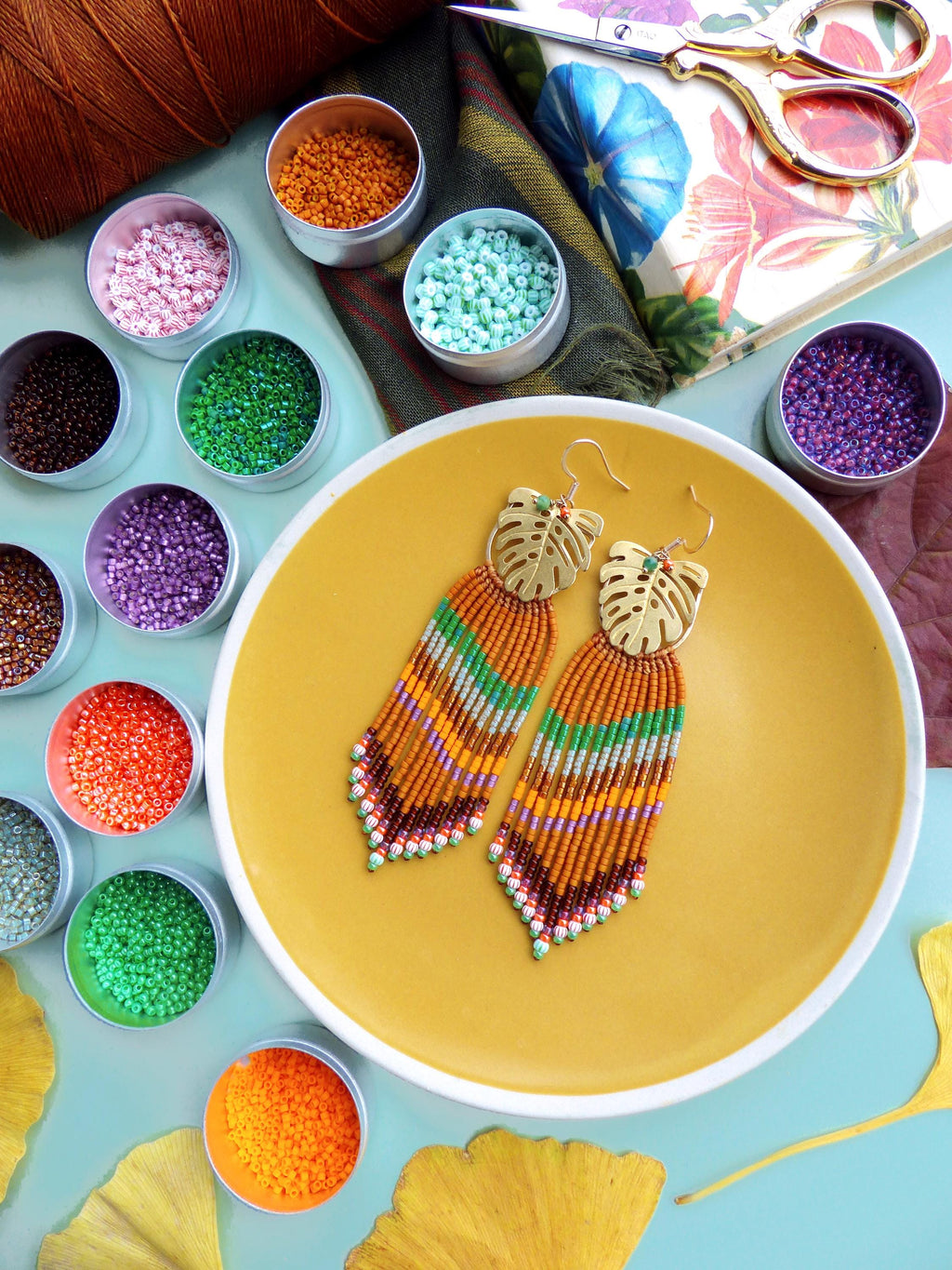Peut inclure: Boucles d'oreilles perlées faites à la main avec des dessus en forme de feuille dorée et une frange de perles orange, vertes et marron. Les boucles d'oreilles reposent sur une assiette jaune, entourées de petits pots de perles colorées et de fournitures d'artisanat.