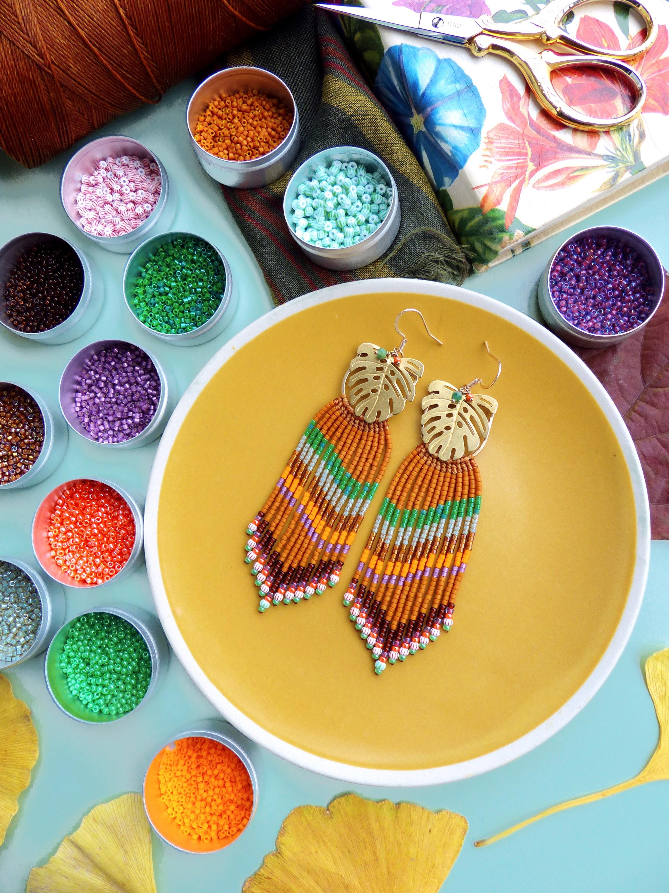 Peut inclure: Boucles d'oreilles perlées faites à la main avec des dessus en forme de feuille dorée et une frange de perles orange, vertes et marron. Les boucles d'oreilles reposent sur une assiette jaune, entourées de petits pots de perles colorées et de fournitures d'artisanat.