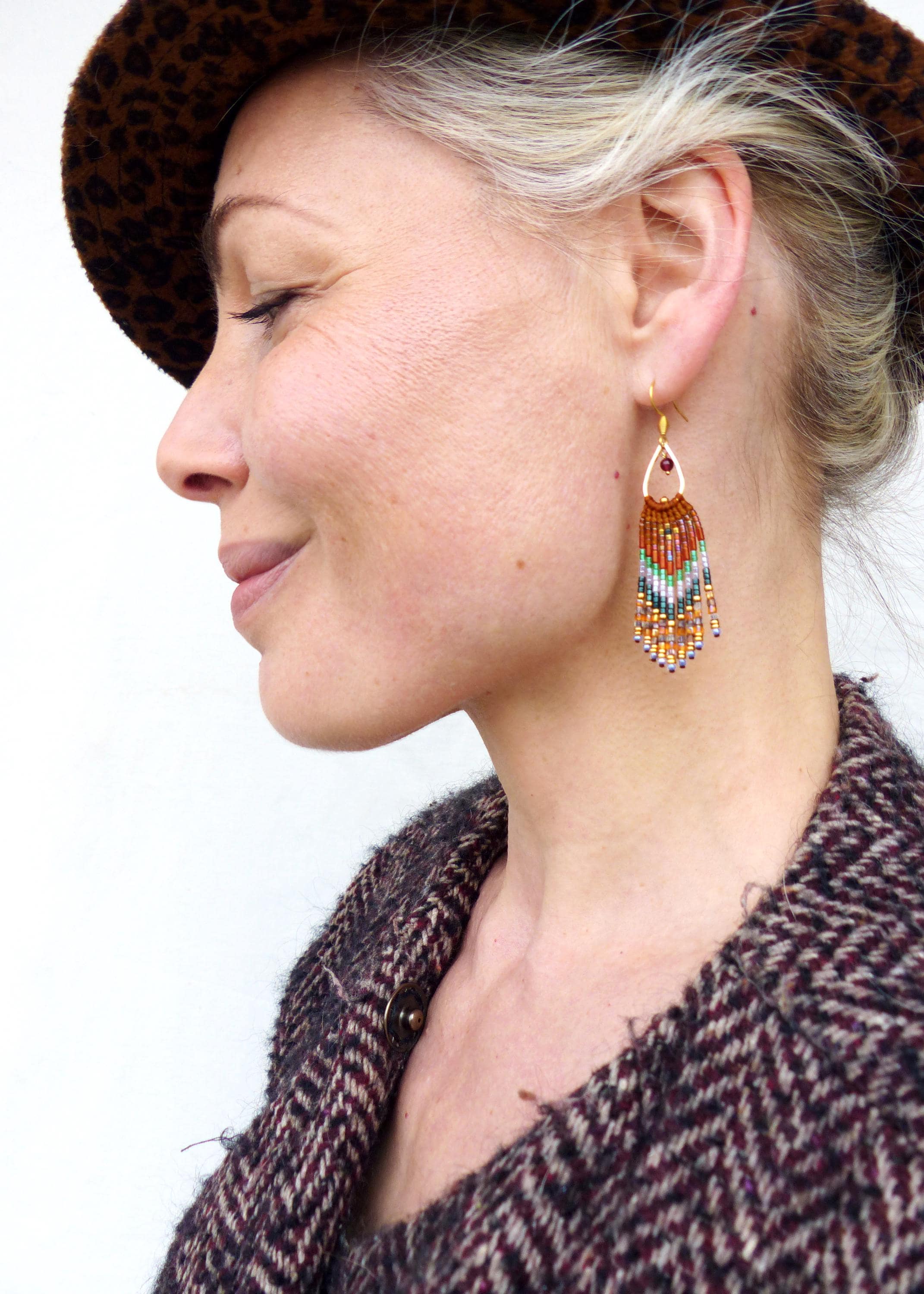 Peut inclure: Une femme portant un chapeau à motif brun et noir et une veste en tweed brun et noir. Elle porte de longues boucles d'oreilles en perles avec un motif géométrique dans des tons d'orange, de vert, de blanc et d'or.