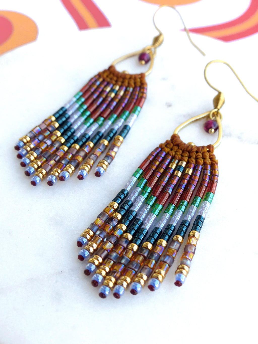 Peut inclure: Une paire de longues boucles d'oreilles pendantes en perles avec un anneau en or et une petite pierre précieuse rouge. Les perles sont dans des tons de brun, rouge, vert, bleu et or.