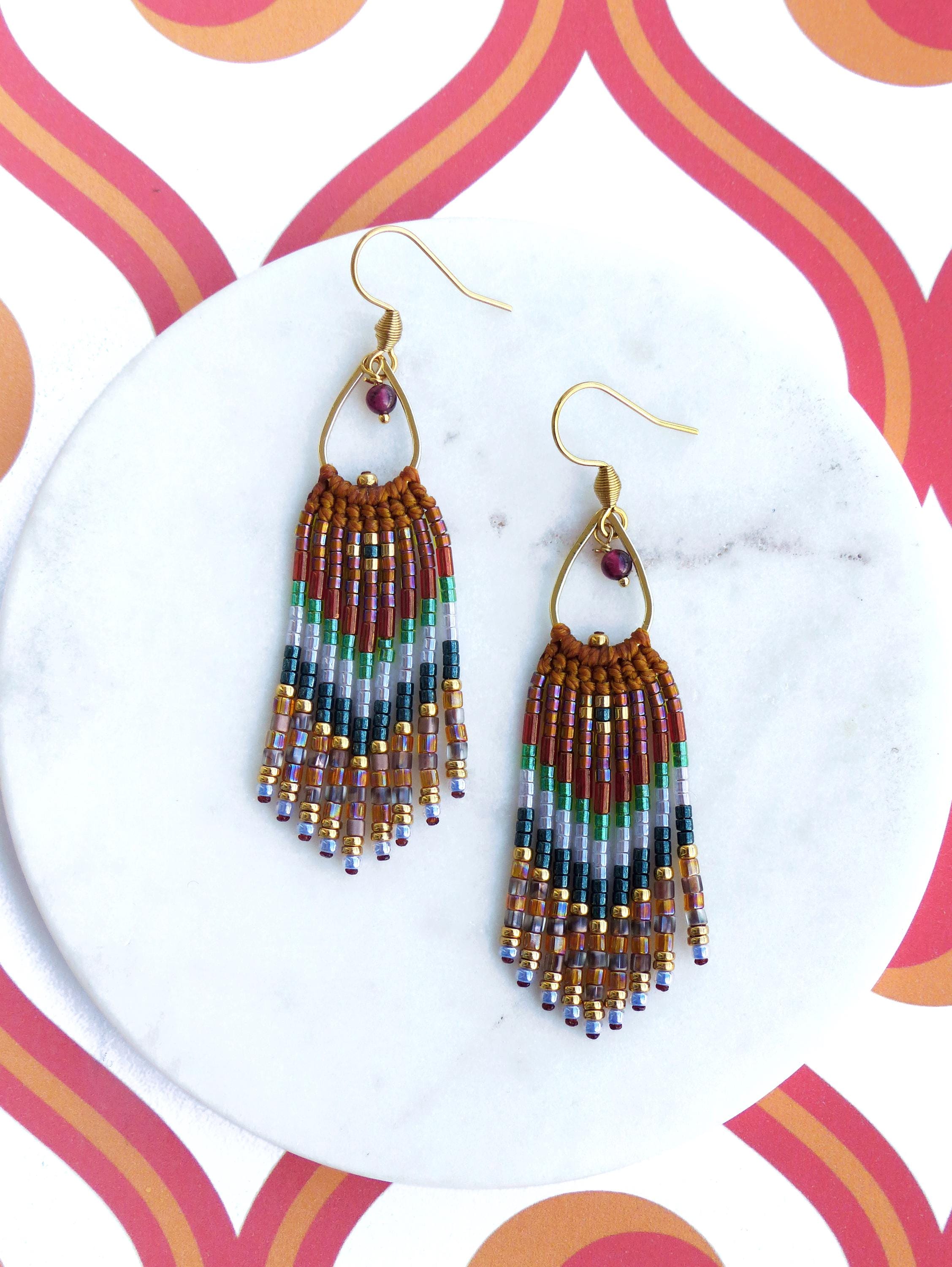 Peut inclure: Une paire de boucles d'oreilles pendantes dorées avec une frange de perles. La frange est composée de perles brunes, vertes, blanches et violettes. Les boucles d'oreilles ont une petite pierre précieuse rouge au sommet.