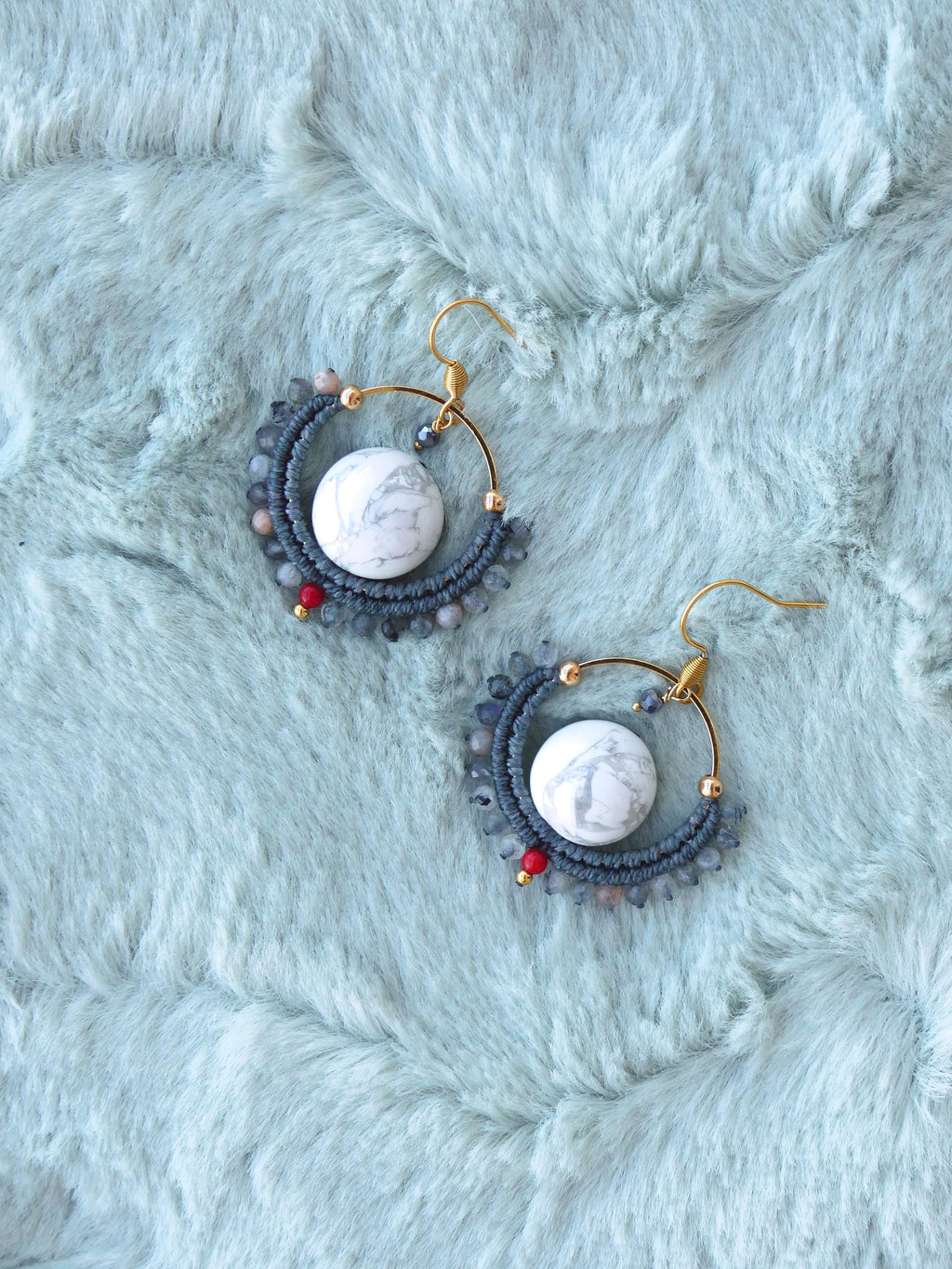 Peut inclure: Boucles d'oreilles artisanales avec un centre en pierre de marbre blanc, entouré d'un motif macramé bleu foncé. Les boucles d'oreilles ont des crochets dorés et sont ornées de petites perles rouges et bleues. Présentées sur une surface duveteuse bleu clair.