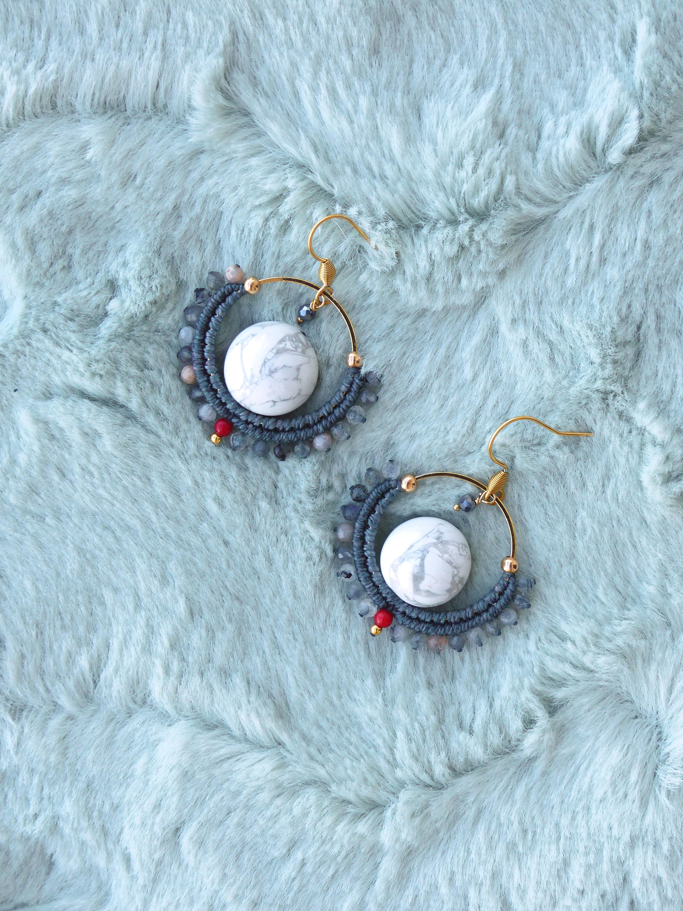 Peut inclure: Boucles d'oreilles artisanales avec un centre en pierre de marbre blanc, entouré d'un motif macramé bleu foncé. Les boucles d'oreilles ont des crochets dorés et sont ornées de petites perles rouges et bleues. Présentées sur une surface duveteuse bleu clair.