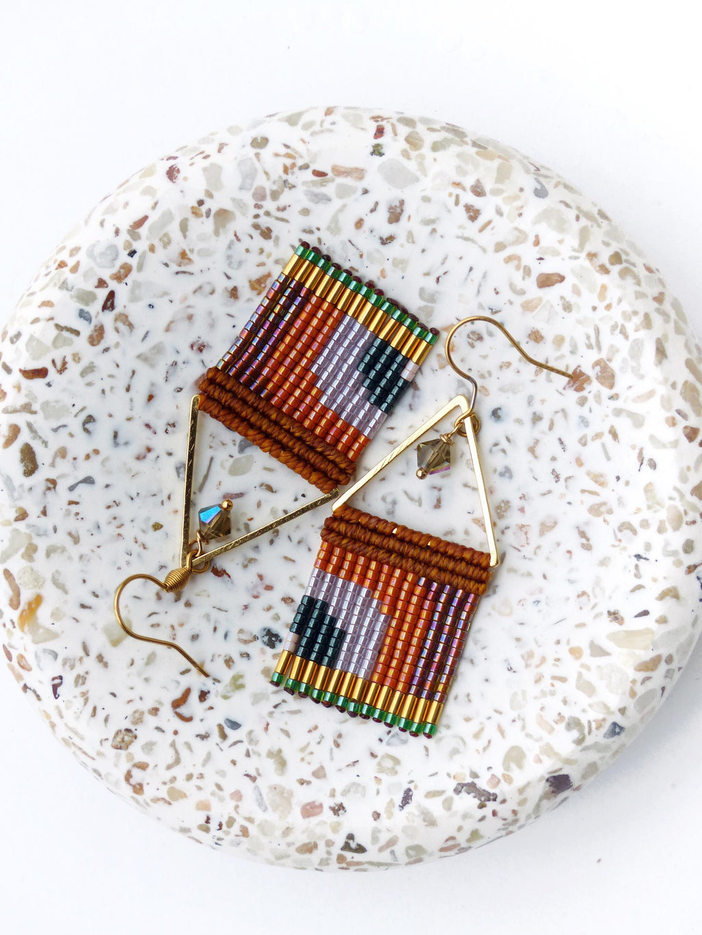 Peut inclure: Une paire de boucles d'oreilles faites à la main avec un motif géométrique. Les boucles d'oreilles présentent un motif tissé dans des tons de marron, orange, violet et vert, suspendu à des cadres triangulaires dorés. Elles sont posées sur une surface blanche mouchetée.